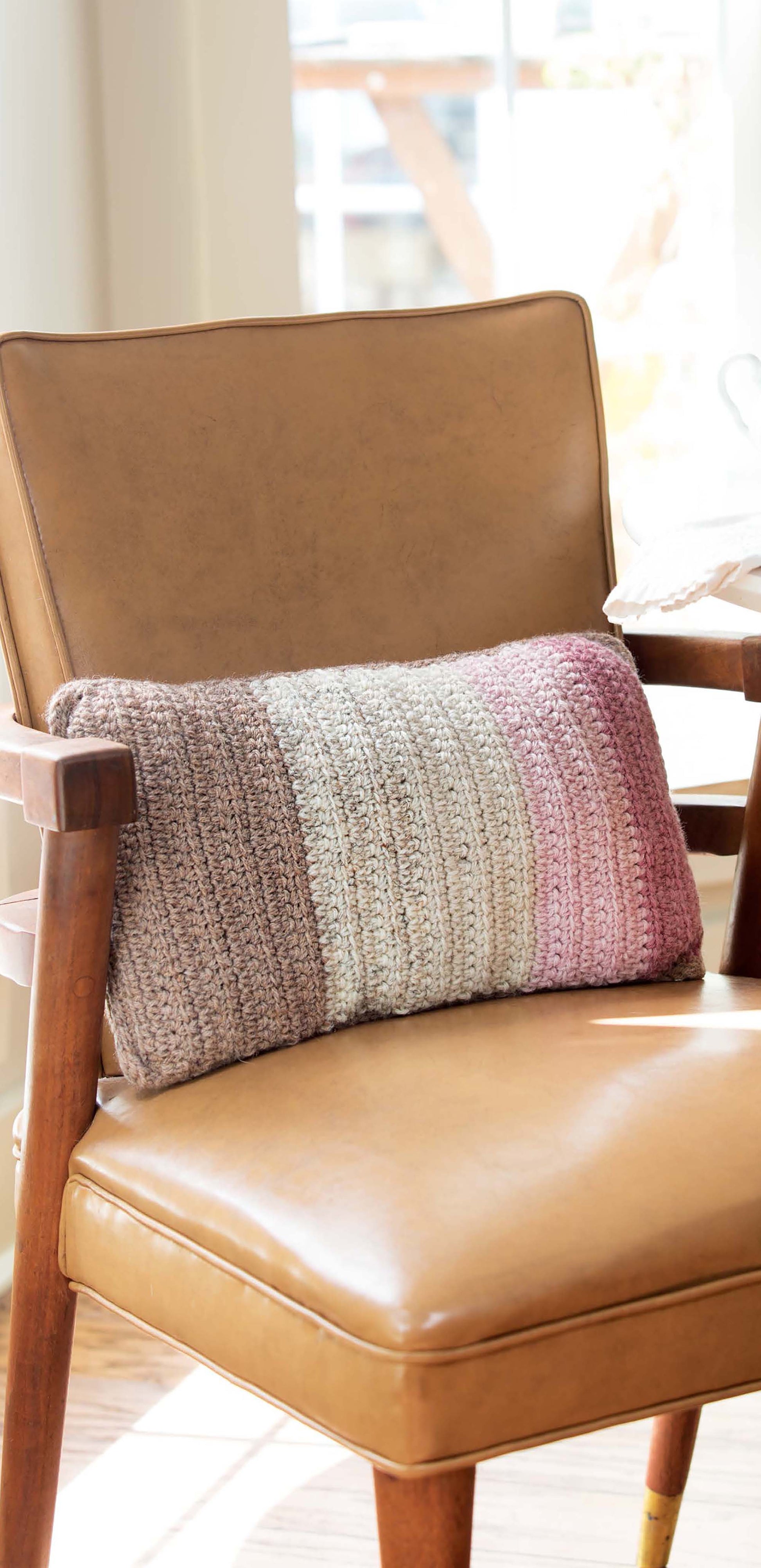 A tan leather armchair with wooden arms holds a rectangular pillow in beige, gray, and pink stripes—an inviting accent inspired by quick projects from "Crochet For The Home" by Leisure Arts—resting in a sunlit room.