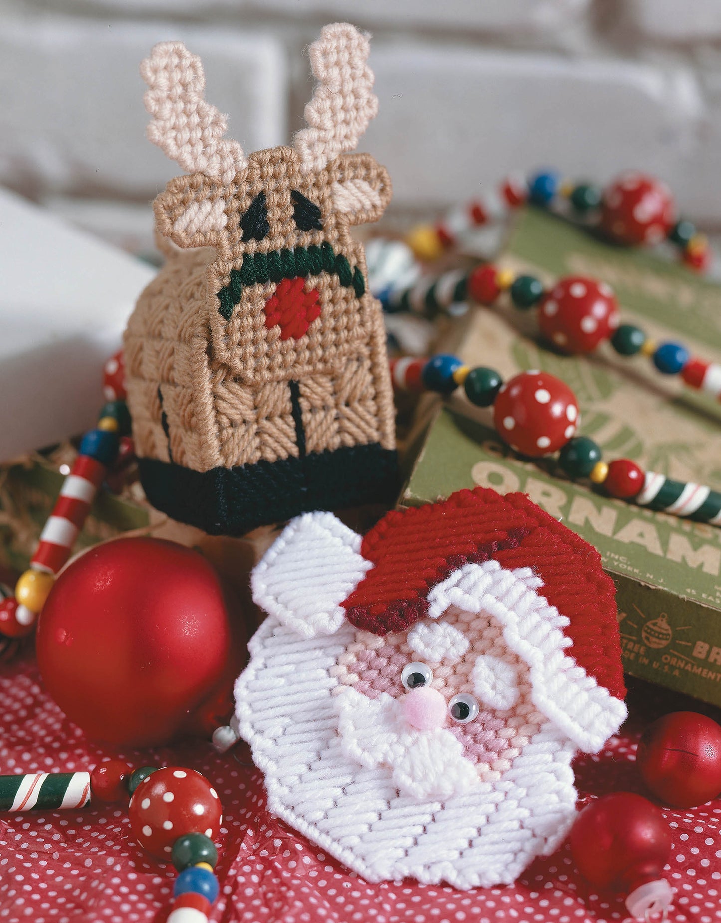 Handmade yarn reindeer and Santa sit among festive plastic canvas ornaments, beads, and boxes in front of a white brick backdrop—inspired by Leisure Arts’ The Magic of Christmas—37 Holiday Plastic Canvas Projects.