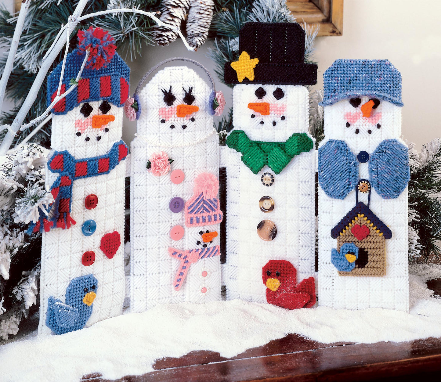 Four handcrafted snowmen from Leisure Arts' "The Magic of Christmas—37 Holiday Plastic Canvas Projects" display unique hats, scarves, and buttons, with embroidered details and small birds by snowy pine branches—perfect for festive crafting.