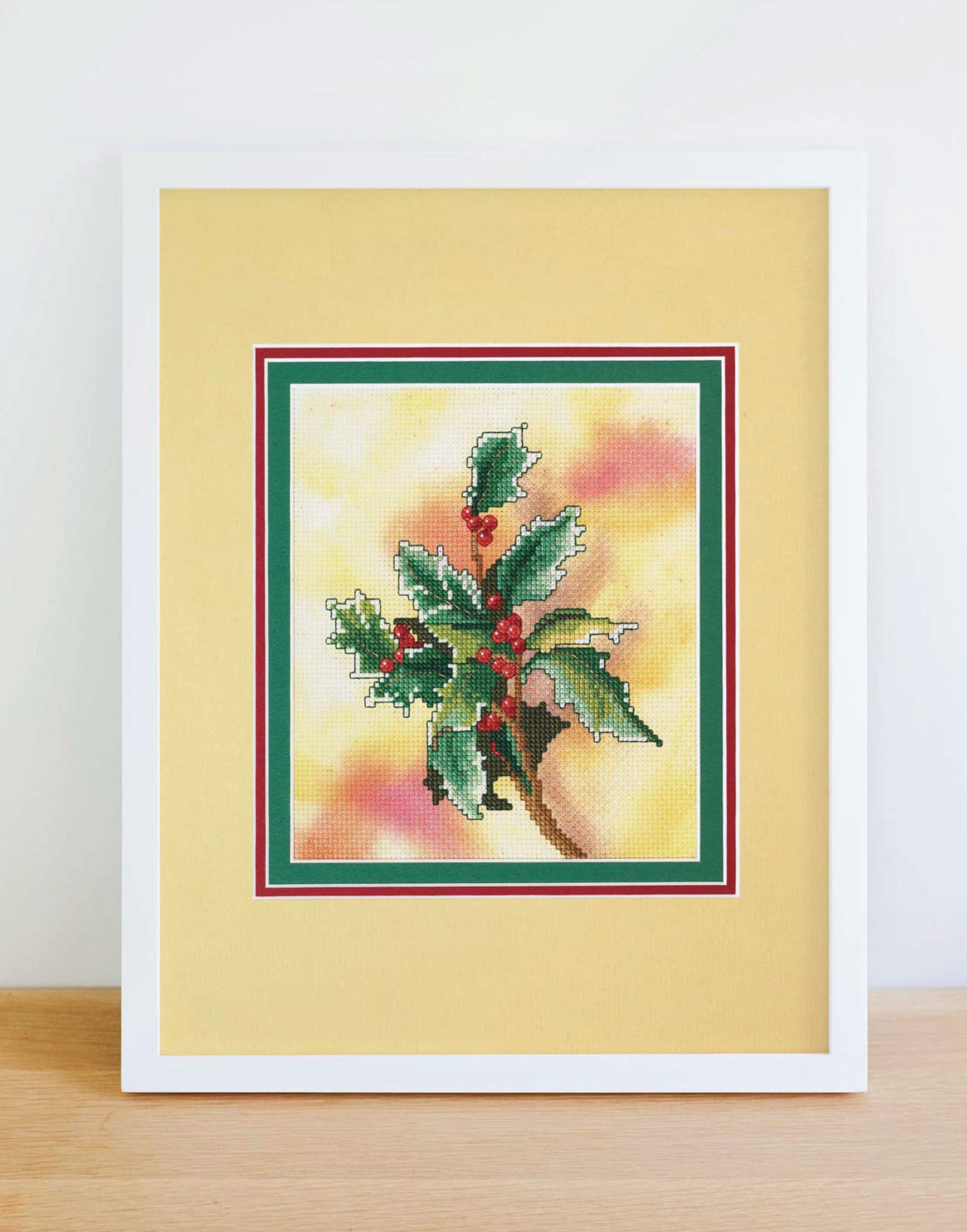 Leisure Arts' Dye-Namic Cross Stitch features green holly leaves and red berries on a colorful background, matted in yellow and displayed on wood—ideal inspiration for your nature-themed cross stitch projects.