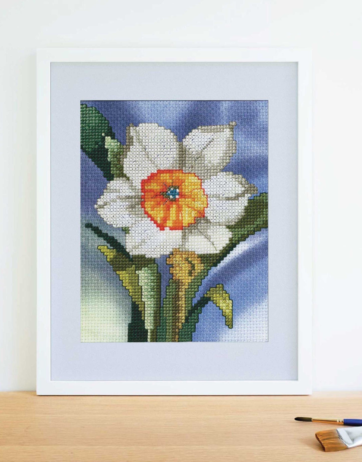 The Leisure Arts Dye-Namic Cross Stitch features a framed white and yellow daffodil with green leaves on a blue and purple background, displayed on wood beside a paintbrush. Inspired by nature designs.