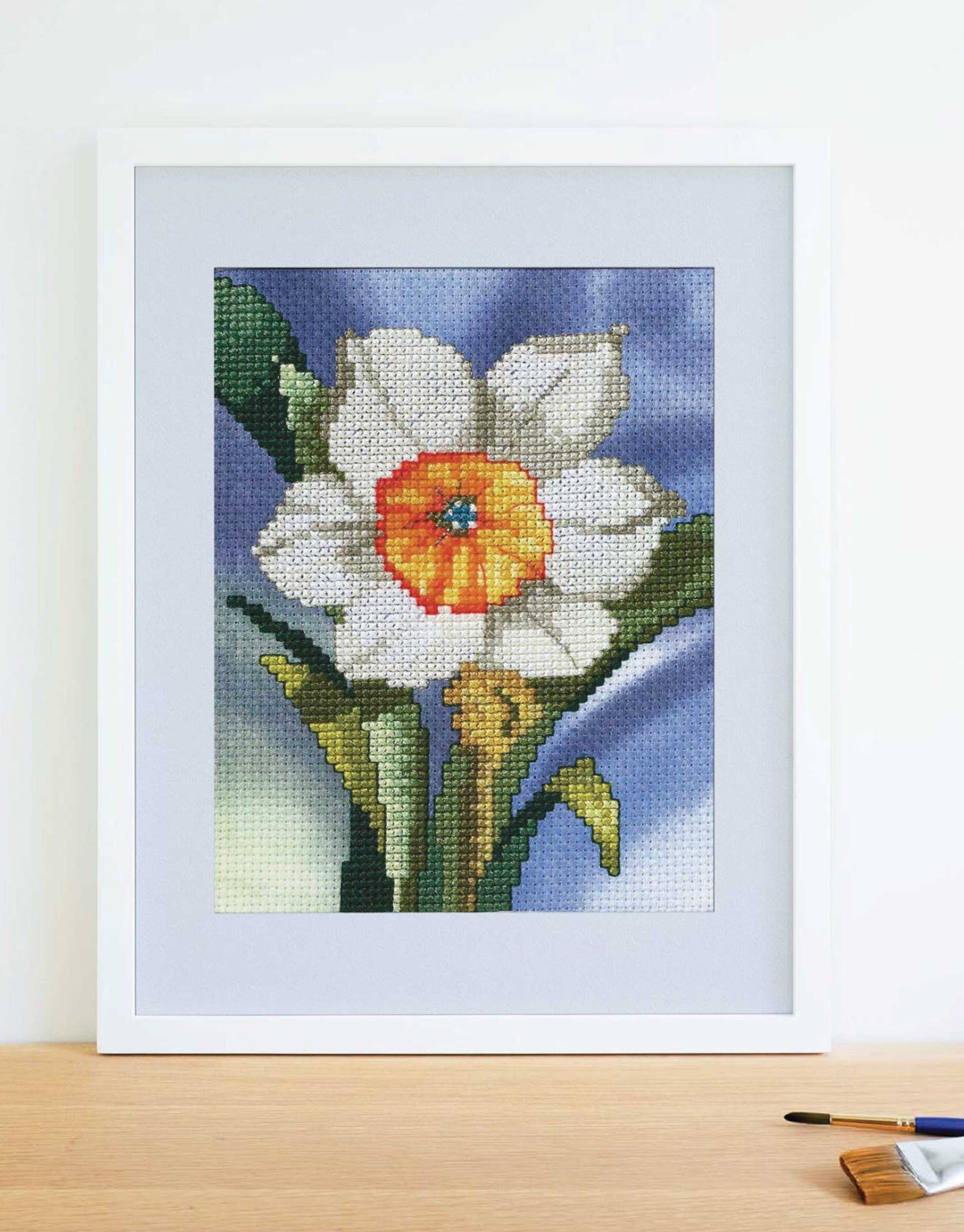 The Leisure Arts Dye-Namic Cross Stitch features a framed white and yellow daffodil with green leaves on a blue and purple background, displayed on wood beside a paintbrush. Inspired by nature designs.