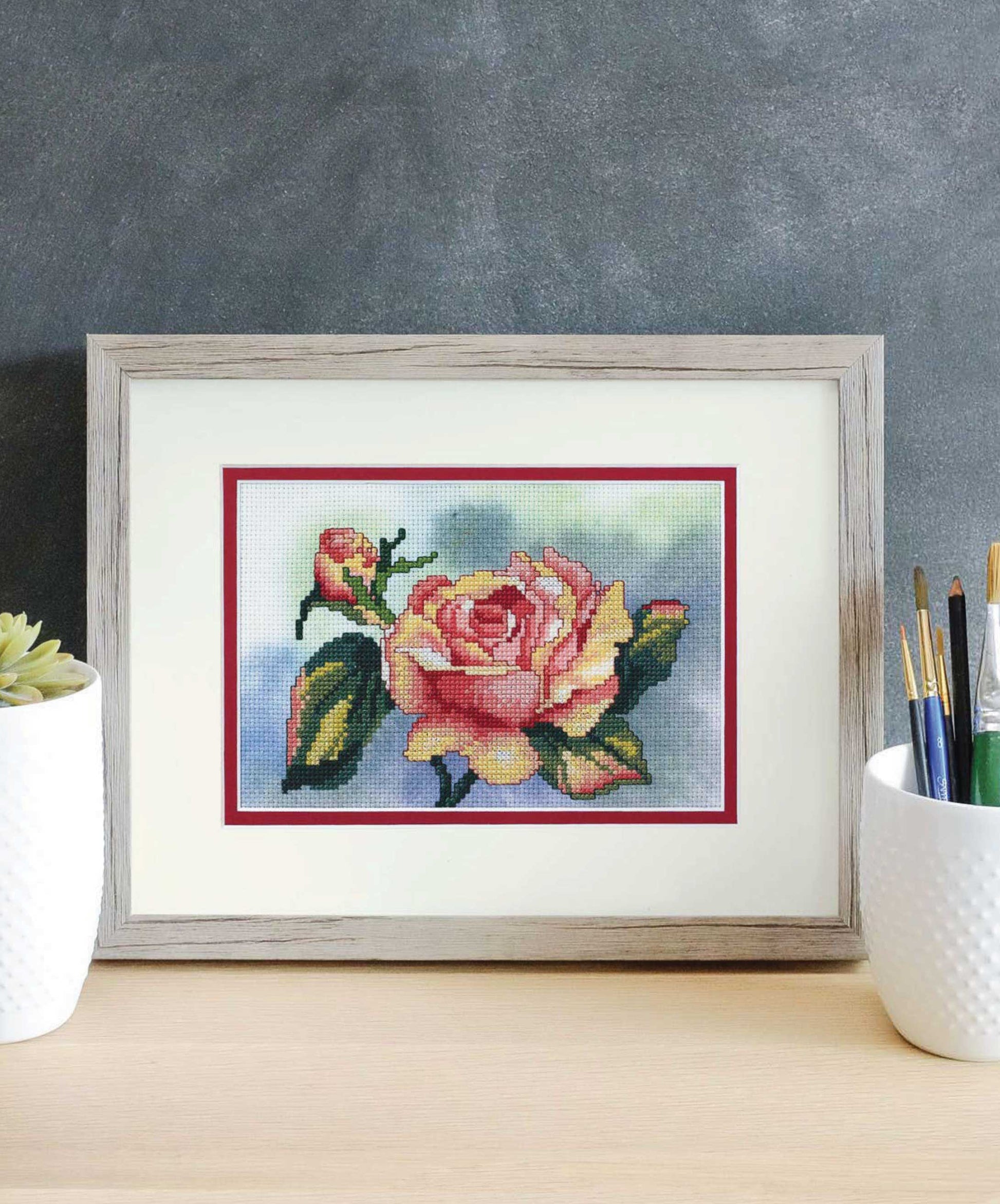 The Dye-Namic Cross Stitch by Leisure Arts, featuring a framed pink and yellow rose with green leaves, is displayed on a wooden desk between a white plant pot and paintbrushes against a dark gray wall.