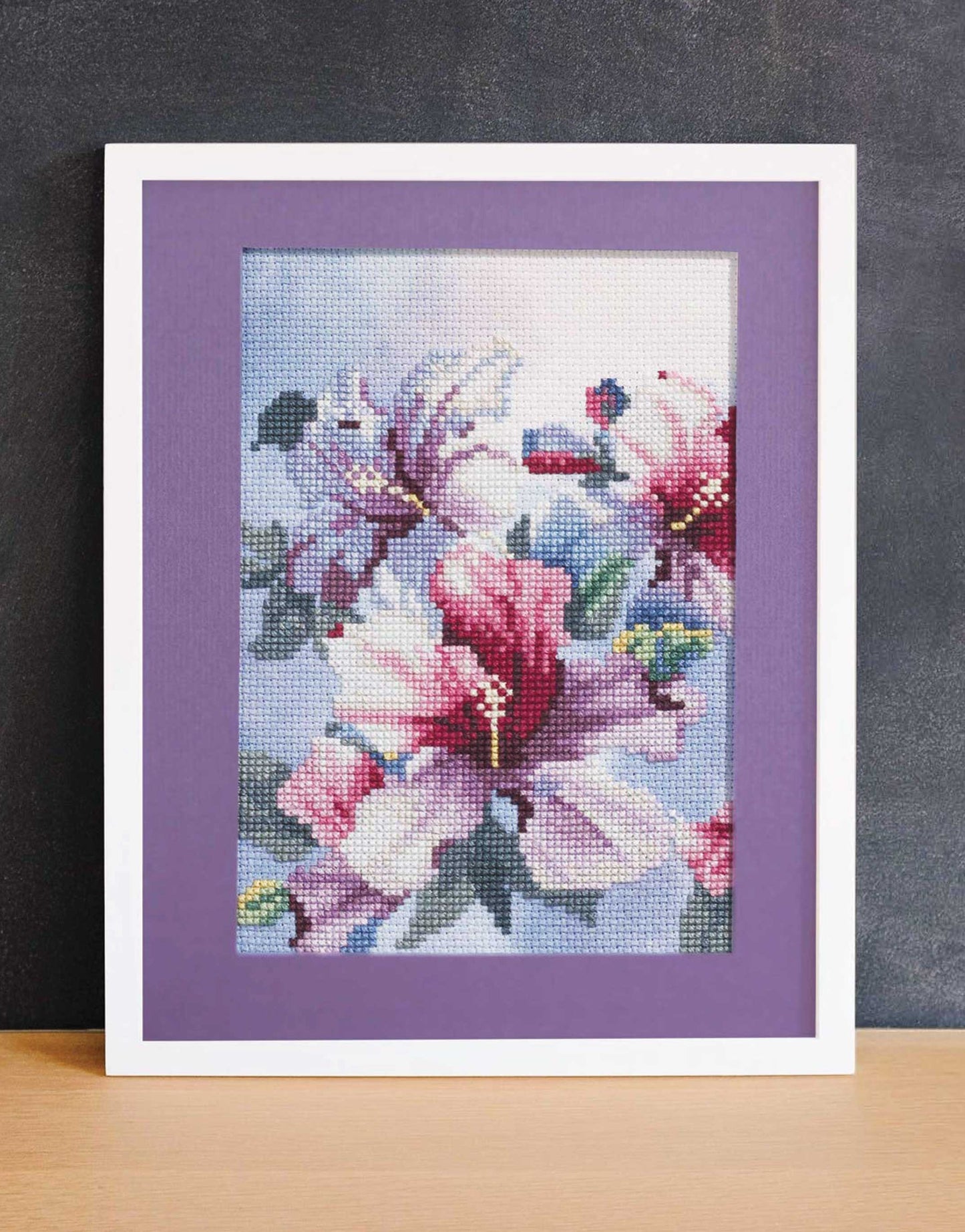 The Dye-Namic Cross Stitch by Leisure Arts features pink and white flowers with green leaves on a soft blue background, bordered by a purple mat—a beautiful way to showcase elegant cross stitch designs in any space.