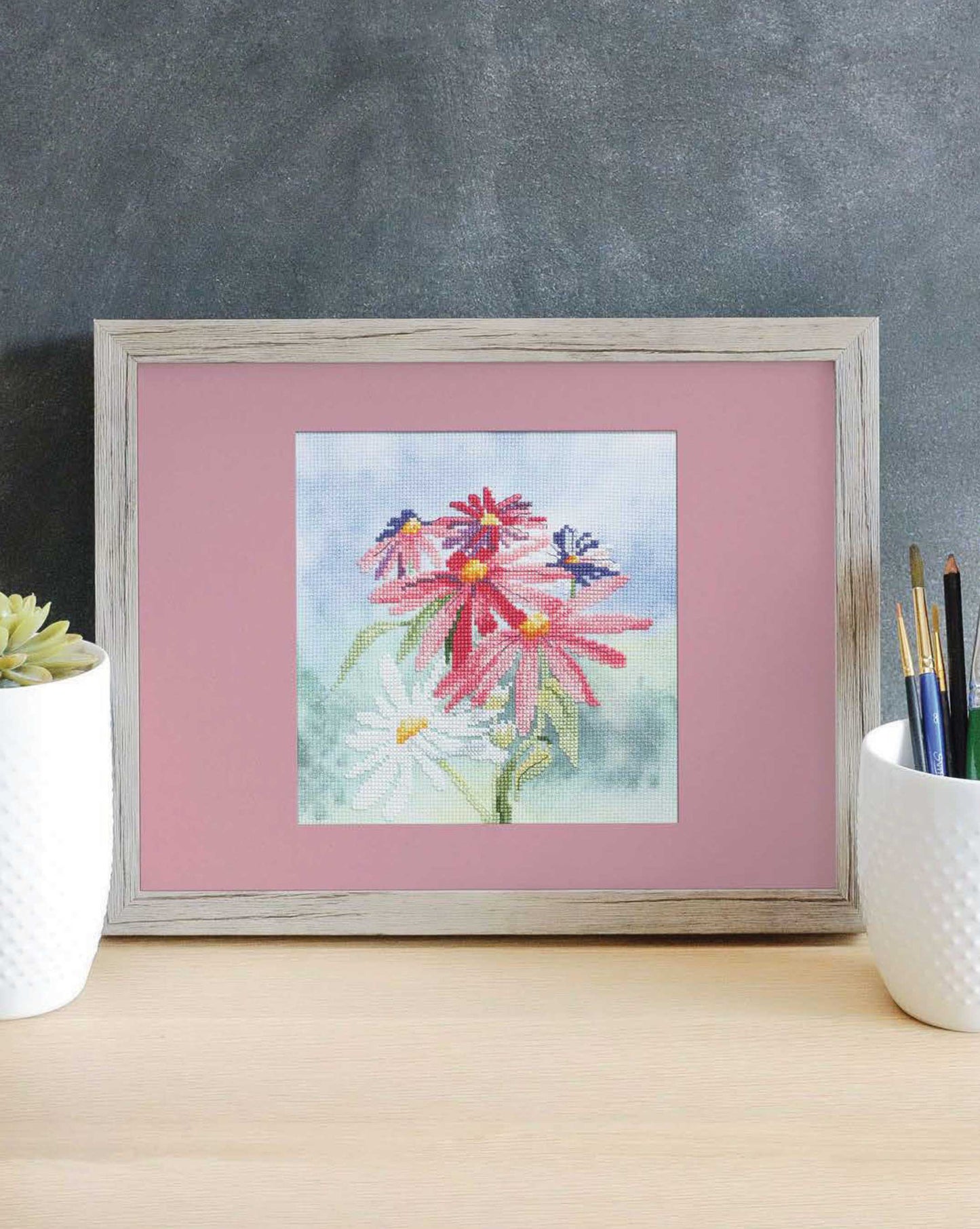 The Dye-Namic Cross Stitch by Leisure Arts features pink and white flowers with green stems on customizable Aida cloth, displayed in a frame on a desk between two white pots—one with a succulent, the other holding paintbrushes.