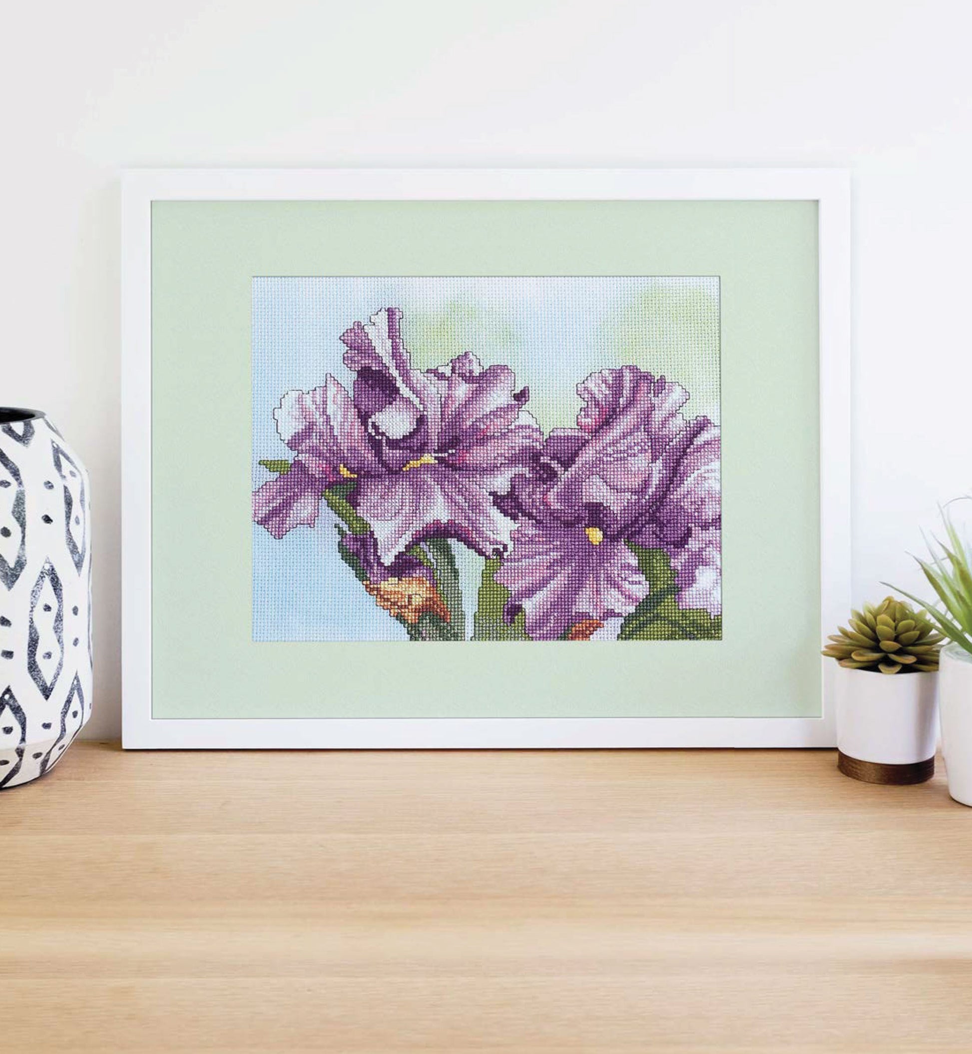 The Leisure Arts Dye-Namic Cross Stitch features two purple irises with green stems, framed and displayed on a light wooden surface between a patterned vase and small potted plants. Ideal for nature lovers or as inspiration for your next stitching project.