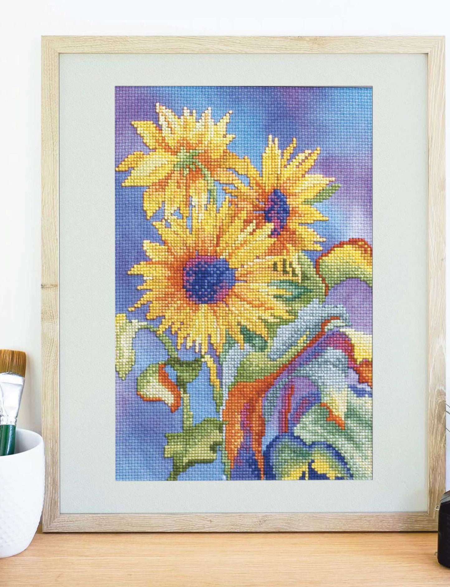 The Dye-Namic Cross Stitch by Leisure Arts features three vibrant yellow sunflowers with green leaves on a purple and blue background, displayed on customizable Aida cloth—perfect for nature lovers—on a light wooden surface.