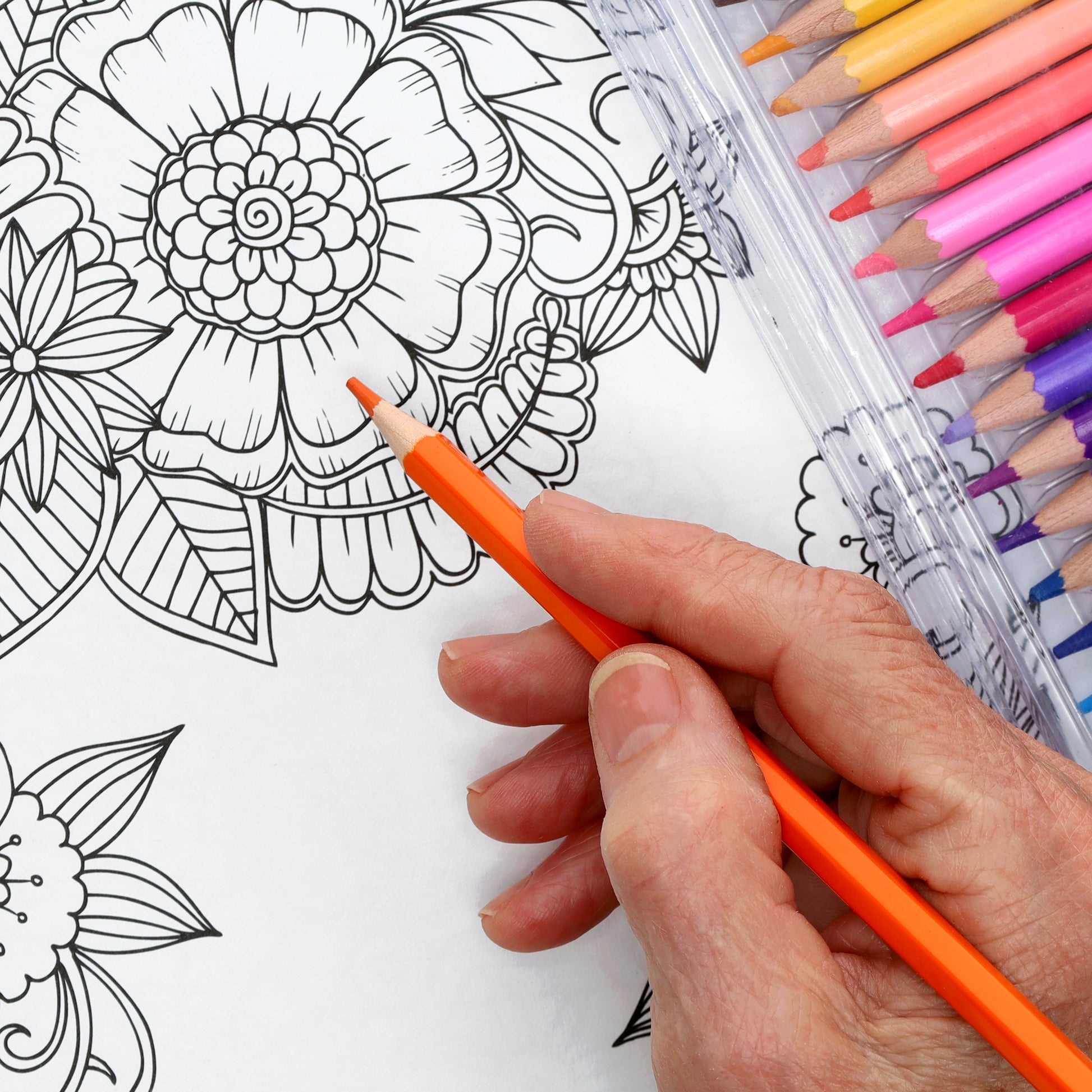 A hand colors a floral page from the Shine On Coloring Book by Leisure Arts using an orange pencil, while pink, purple, and orange pencils lie nearby—ideal inspiration for your 32 creative designs.
