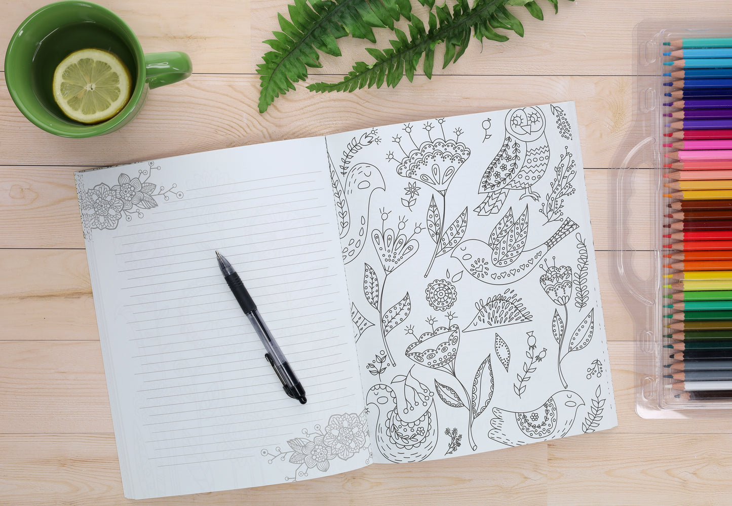 An open notebook displays lined pages with a pen and the Leisure Arts Shine On Coloring Book page featuring birds and flowers. To the right are colored pencils, above is a green cup with lemon, and a fern leaf rests on the wooden surface.