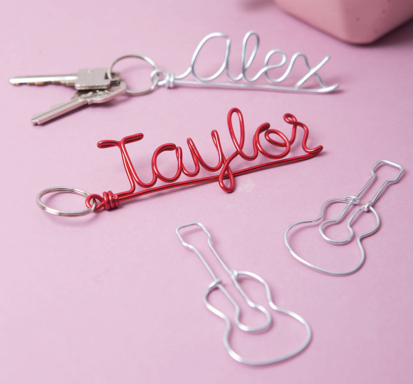 Several wire keychains crafted from art wire rest on a pink surface, inspired by projects from Leisure Arts' "Wire Crafts—18 Wire Wrapping Projects to Bend, Wrap and Twist." Partially visible are a set of keys and a pink object.