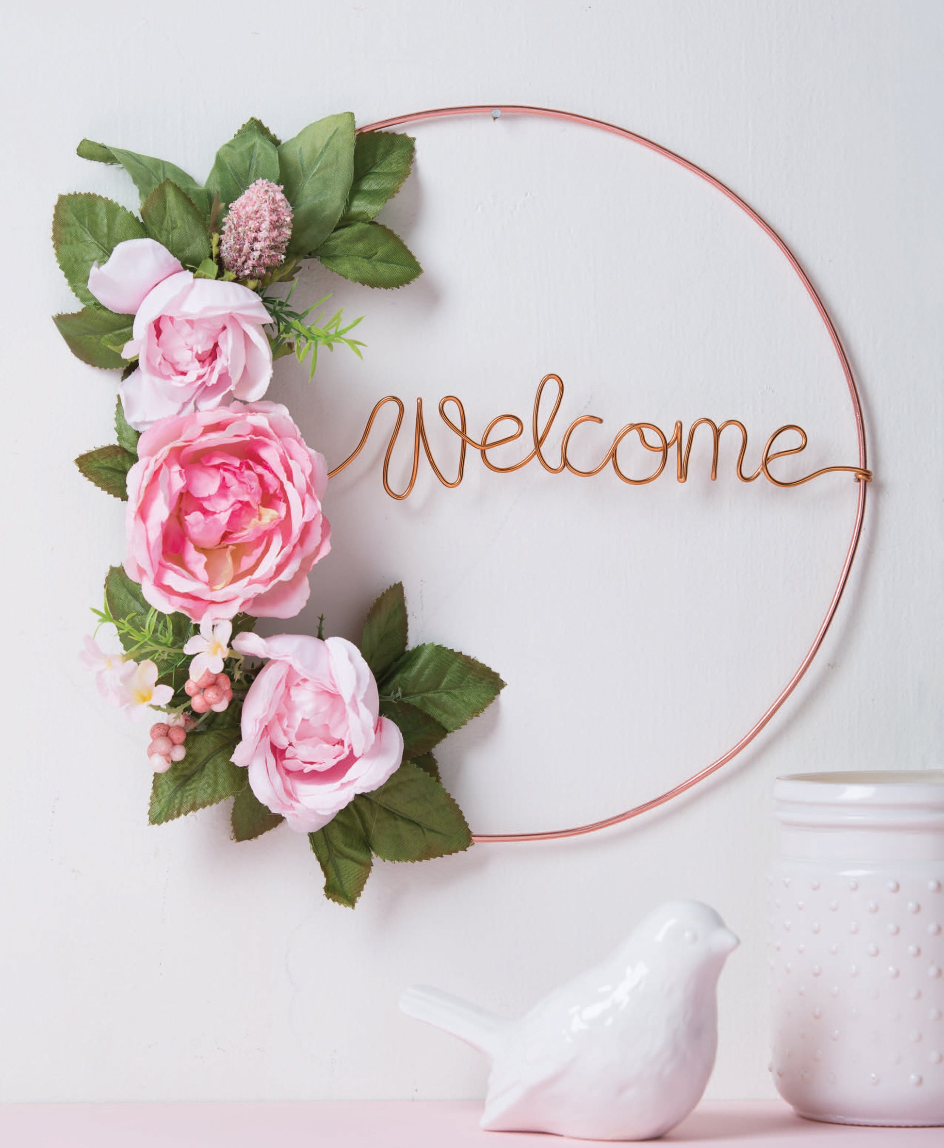 Wire Crafts—18 Wire Wrapping Projects to Bend, Wrap and Twist by Leisure Arts features inspiring designs like a pink flower and green leaf wreath with a wire "Welcome" sign—perfect for beginner home decor or easy projects. Ceramic bird and jar accents shown.