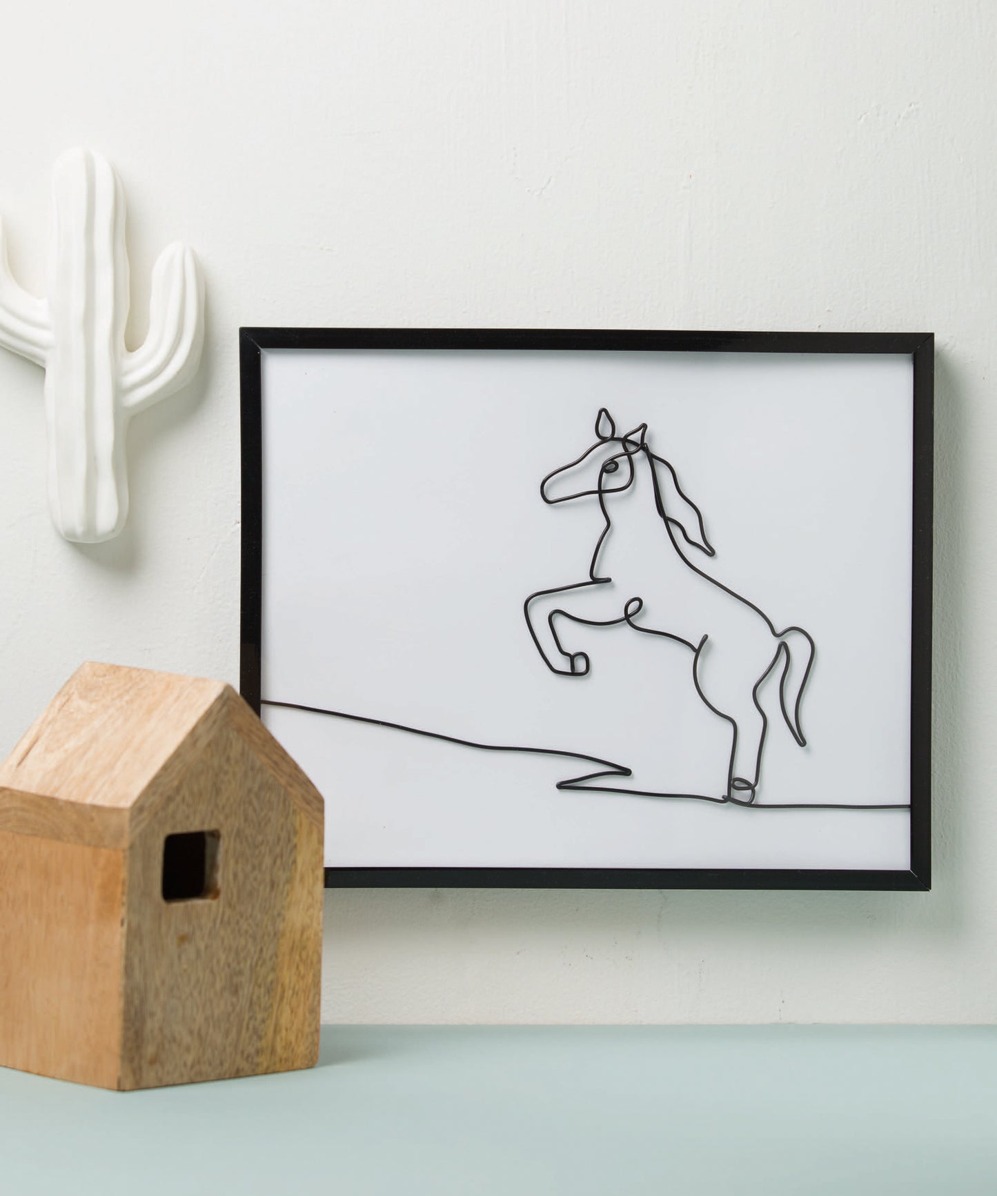 Minimalist decor scene with a framed art wire drawing of a prancing horse from "Wire Crafts—18 Wire Wrapping Projects to Bend, Wrap and Twist" by Leisure Arts, displayed with a small wooden house ornament and a white ceramic cactus.