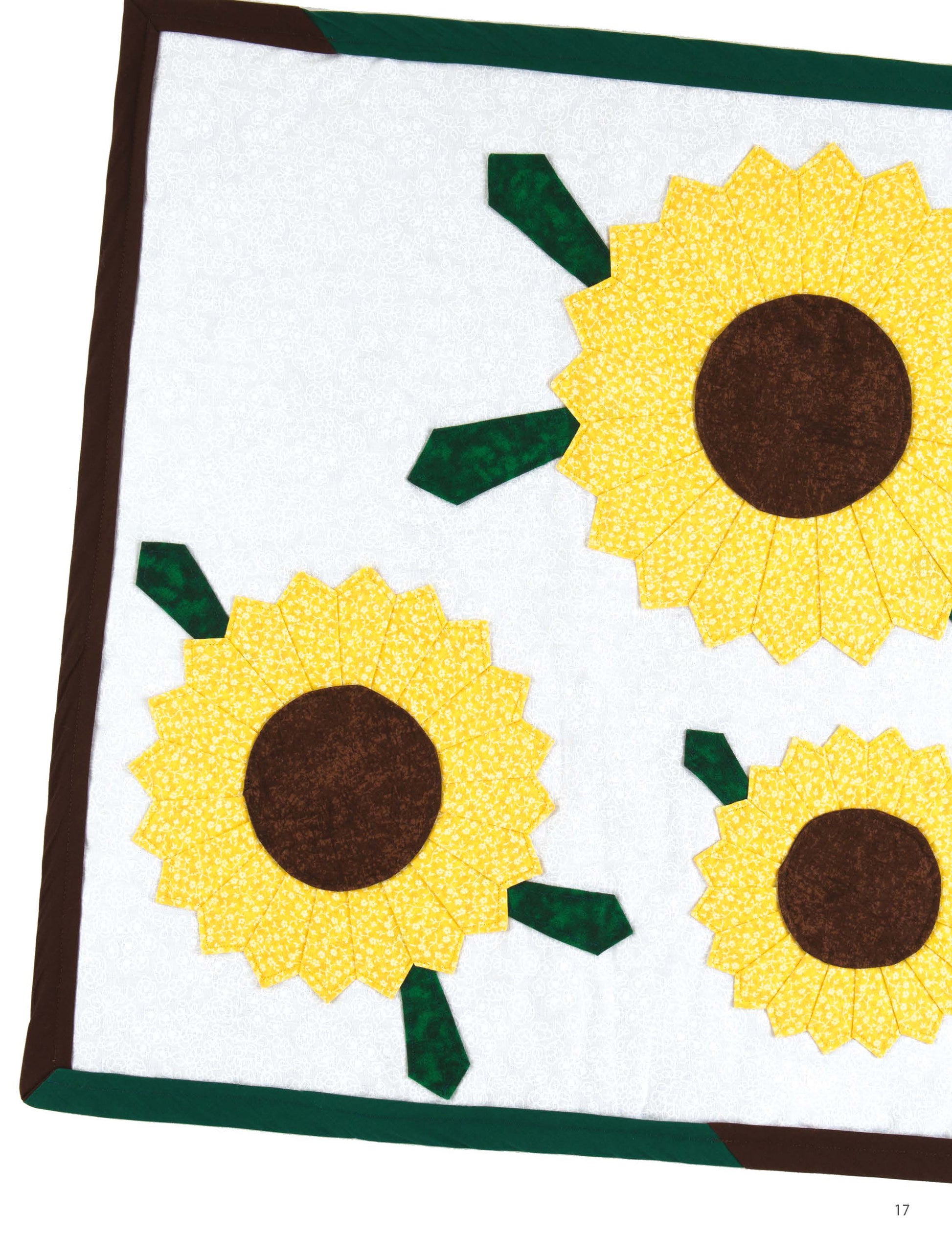 A charming mini quilt from Leisure Arts’ "Miniature Quilts—12 Tiny Projects That Make a Big Impression" features three yellow sunflowers with brown centers on white, bordered in brown. The number 17 appears at the bottom right corner. Perfect for home decor gifts!.