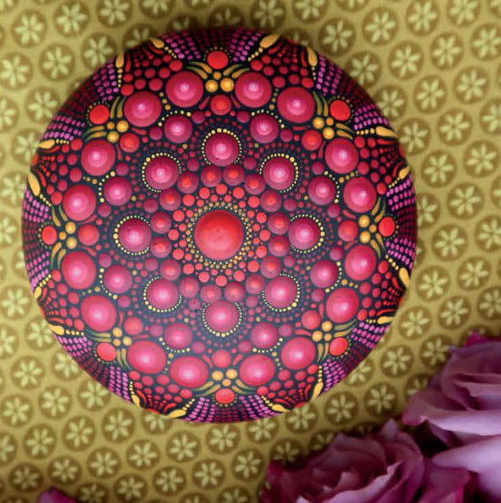 The Dot Painting Digital Download by Leisure Arts features a round mandala in red, pink, orange, and yellow dots displayed on a gold floral-patterned surface with pink rose petals in the lower right corner.