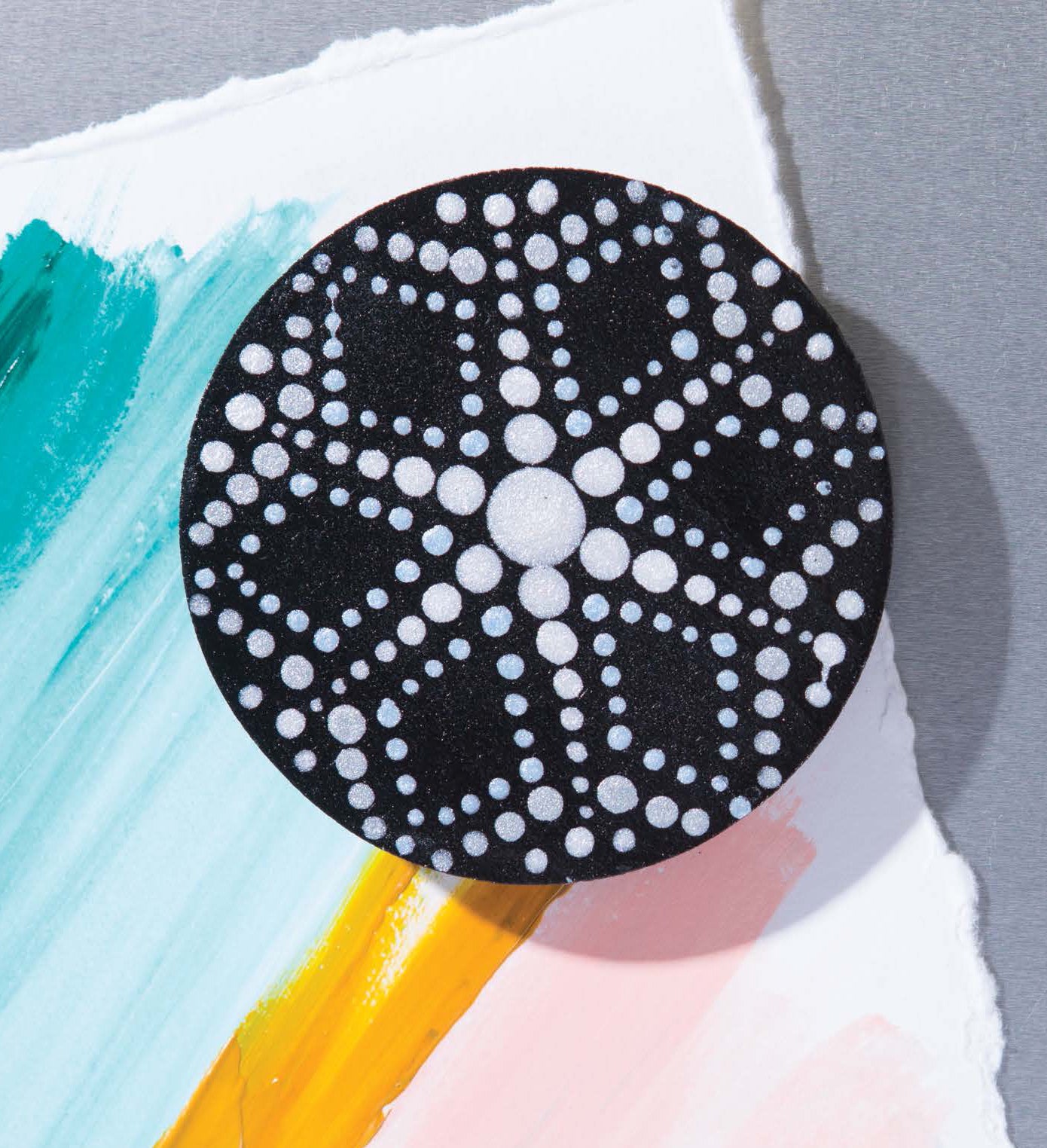 The Dot Painting Digital Download by Leisure Arts features a round black design with white geometric dot patterns, displayed on textured white paper with teal, yellow, and peach watercolor brushstrokes against a gray background.