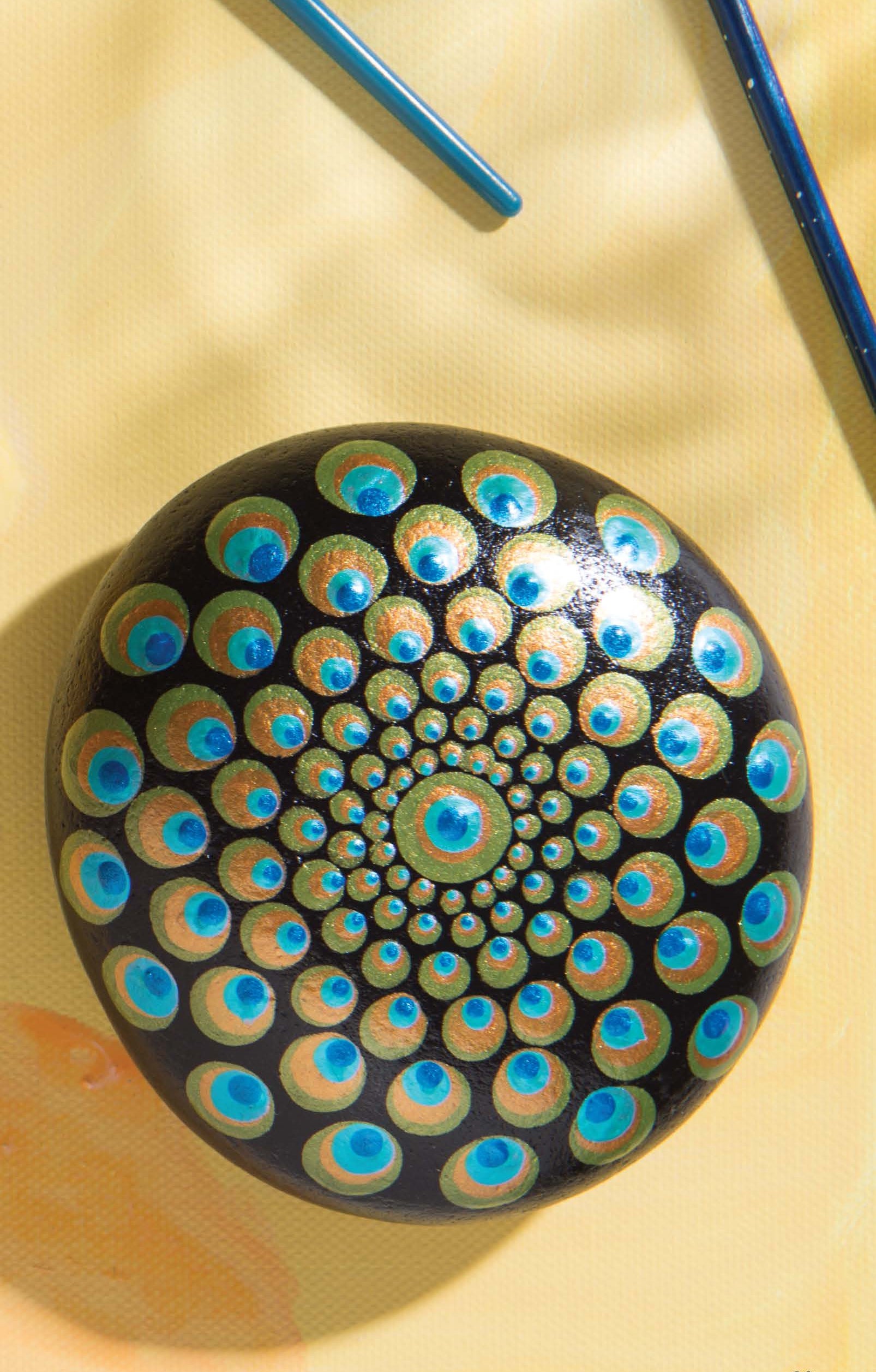 Leisure Arts' Dot Painting Digital Download features a round black stone adorned with a vibrant mandala of gold, blue, and turquoise dots. Paintbrushes rest nearby on a light yellow surface, showcasing creative rock painting techniques.