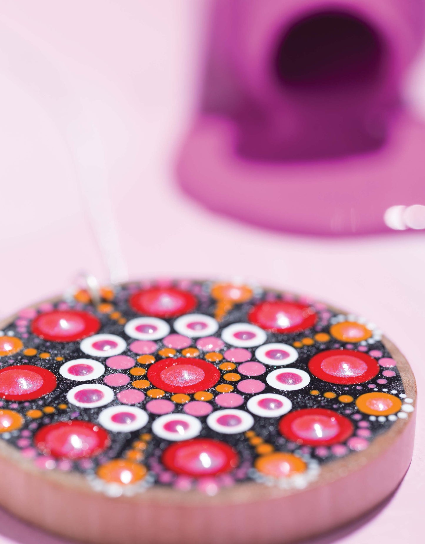 The Dot Painting Digital Download by Leisure Arts features a round mandala design in red, orange, pink, black, and white resting on a pink surface with a blurred purple object in the background.