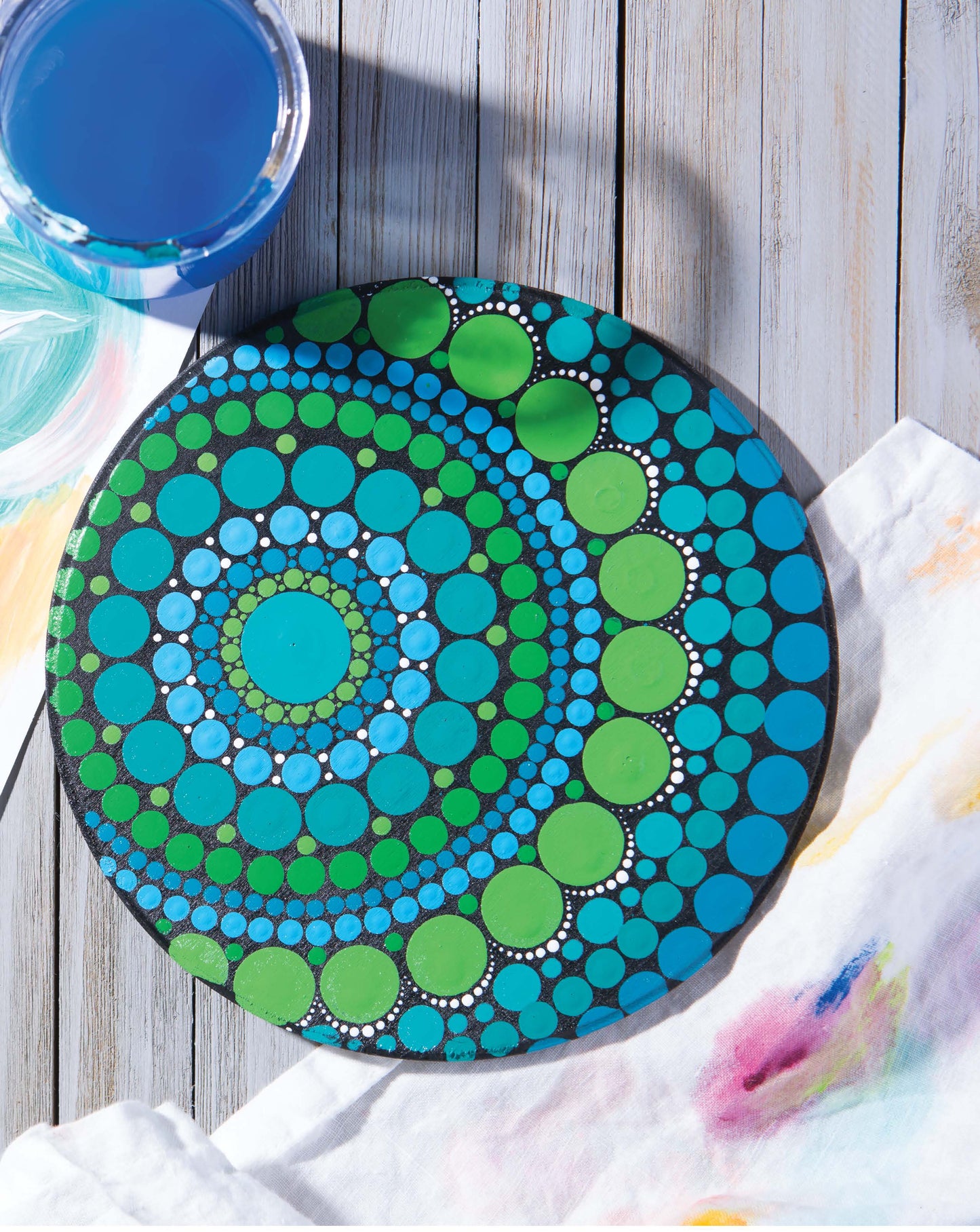 The Dot Painting Digital Download by Leisure Arts features a black round stone with vivid blue and green mandala art, displayed on a light wooden surface beside blue water and a paint-stained white cloth.