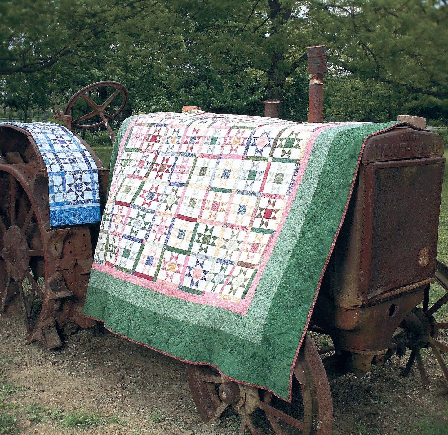 Two quilts made with triangle piecing from Leisure Arts’ It's Quilt Magic Digital Download are draped over a vintage tractor outdoors, with green grass and trees in the background.