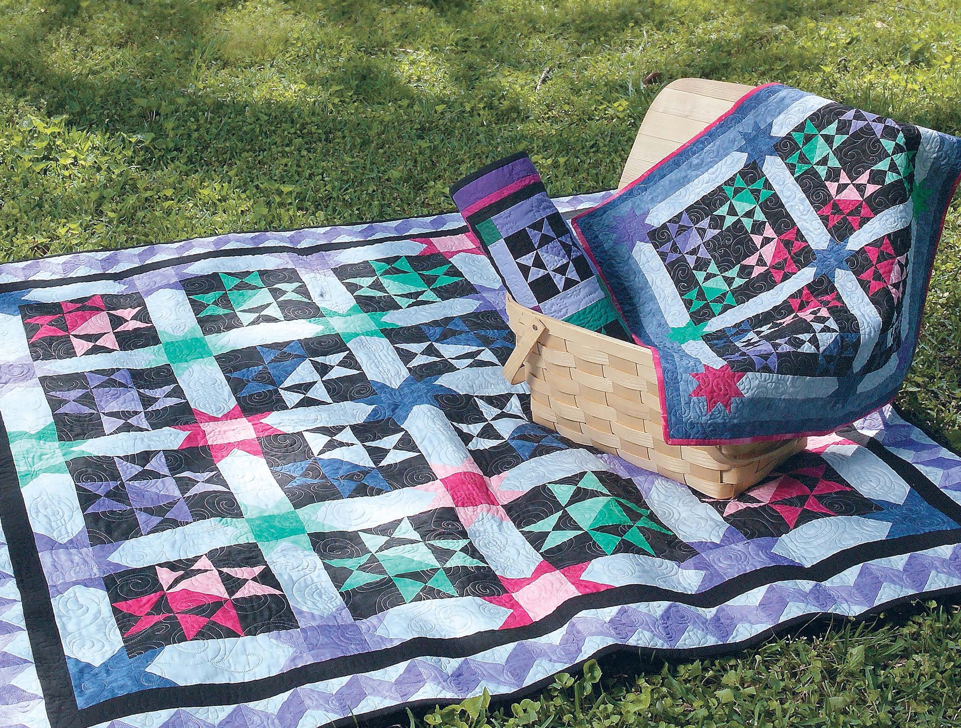 The “It’s Quilt Magic Digital Download” by Leisure Arts features a vibrant quilt with geometric patterns, displayed on green grass beside a woven basket holding a matching quilt and intricately pieced triangle fabric.
