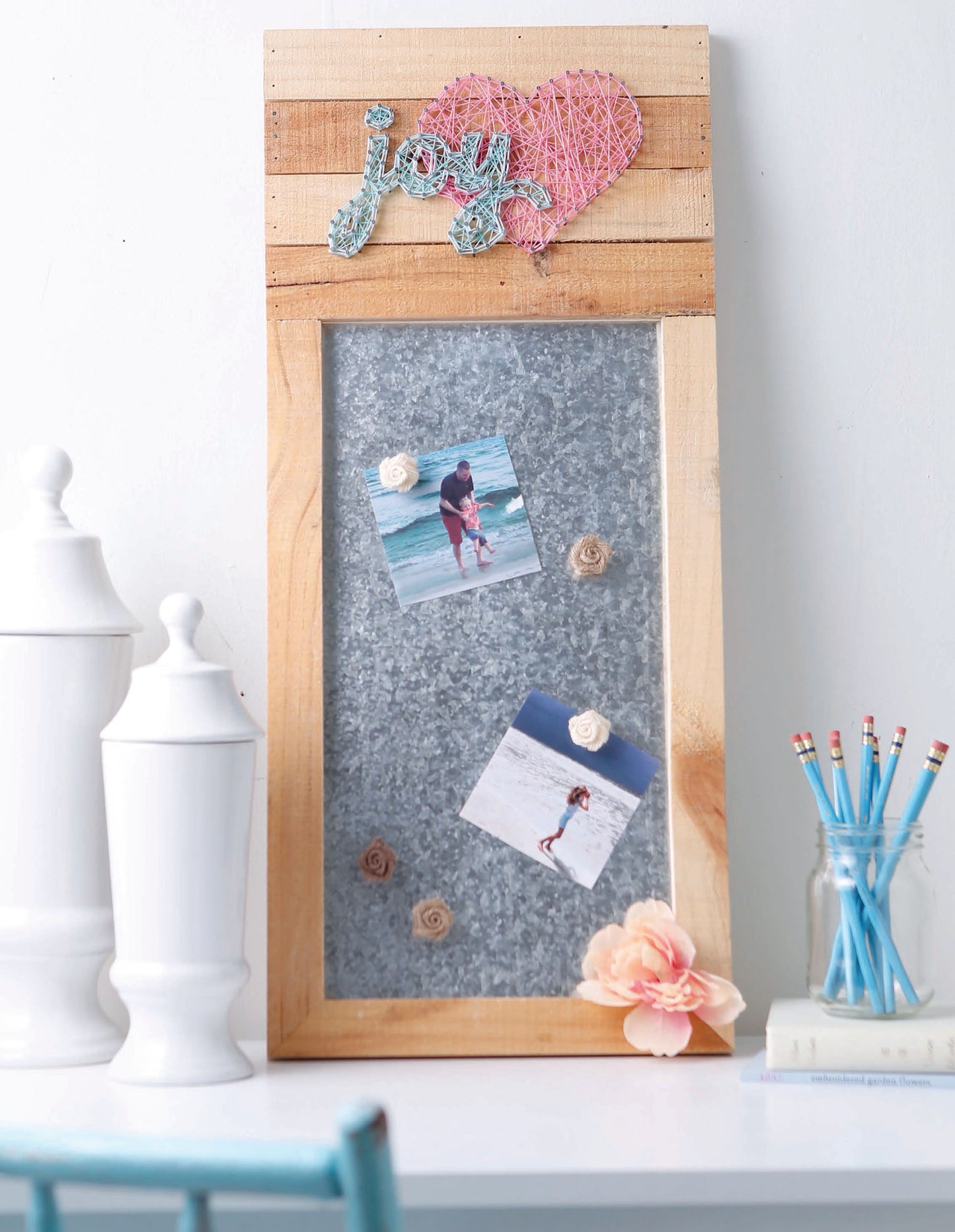 The Leisure Arts String Art 2 memo board features “joy” and a heart in wooden and metal string art, displays two beach photos and flower magnets, and is shown on a table with white jars, a bouquet, and blue pencils.