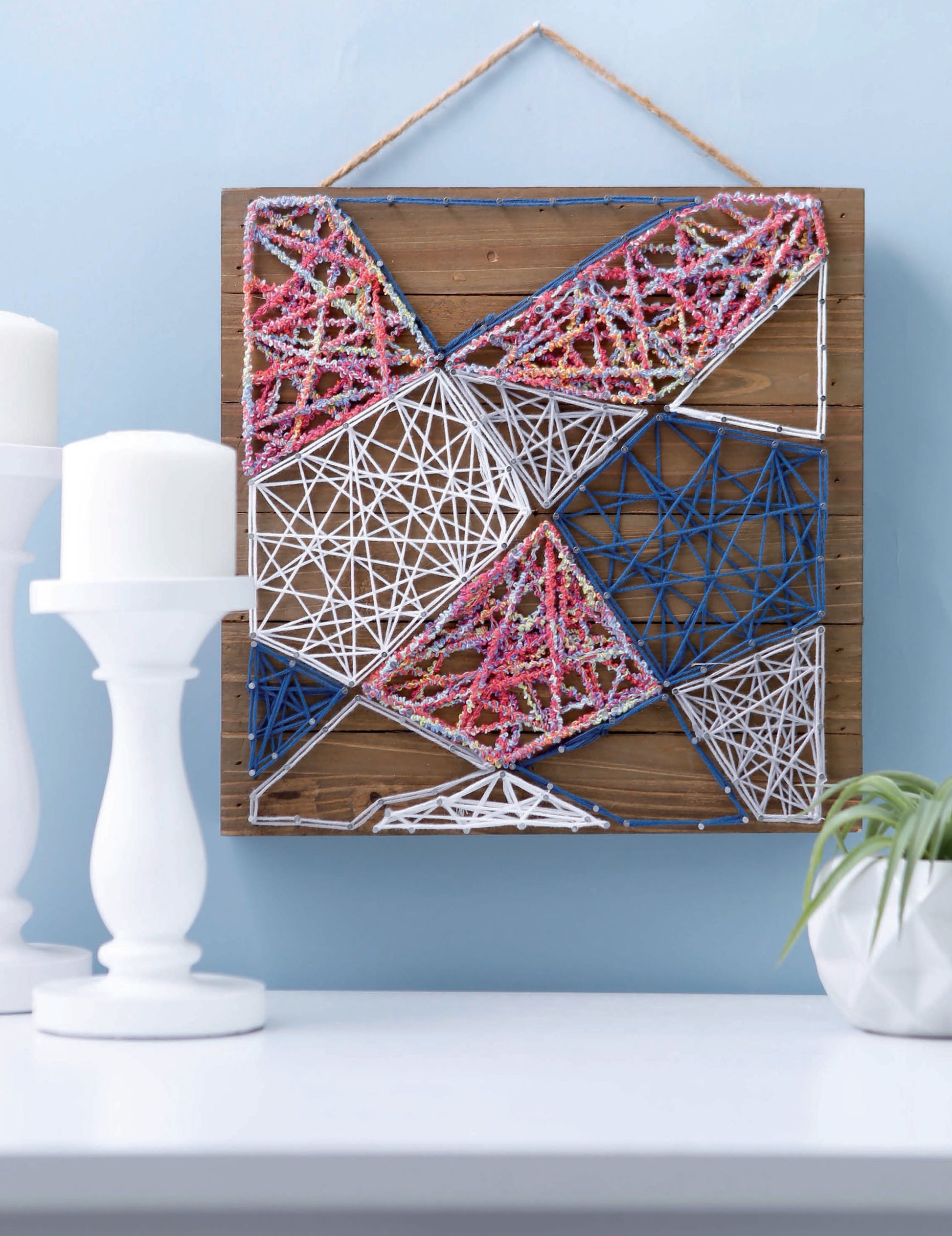 Leisure Arts’ String Art 2 features blue, white, and pink threads on a wooden board, displayed on a light blue wall above a white table with candles and a small green plant.