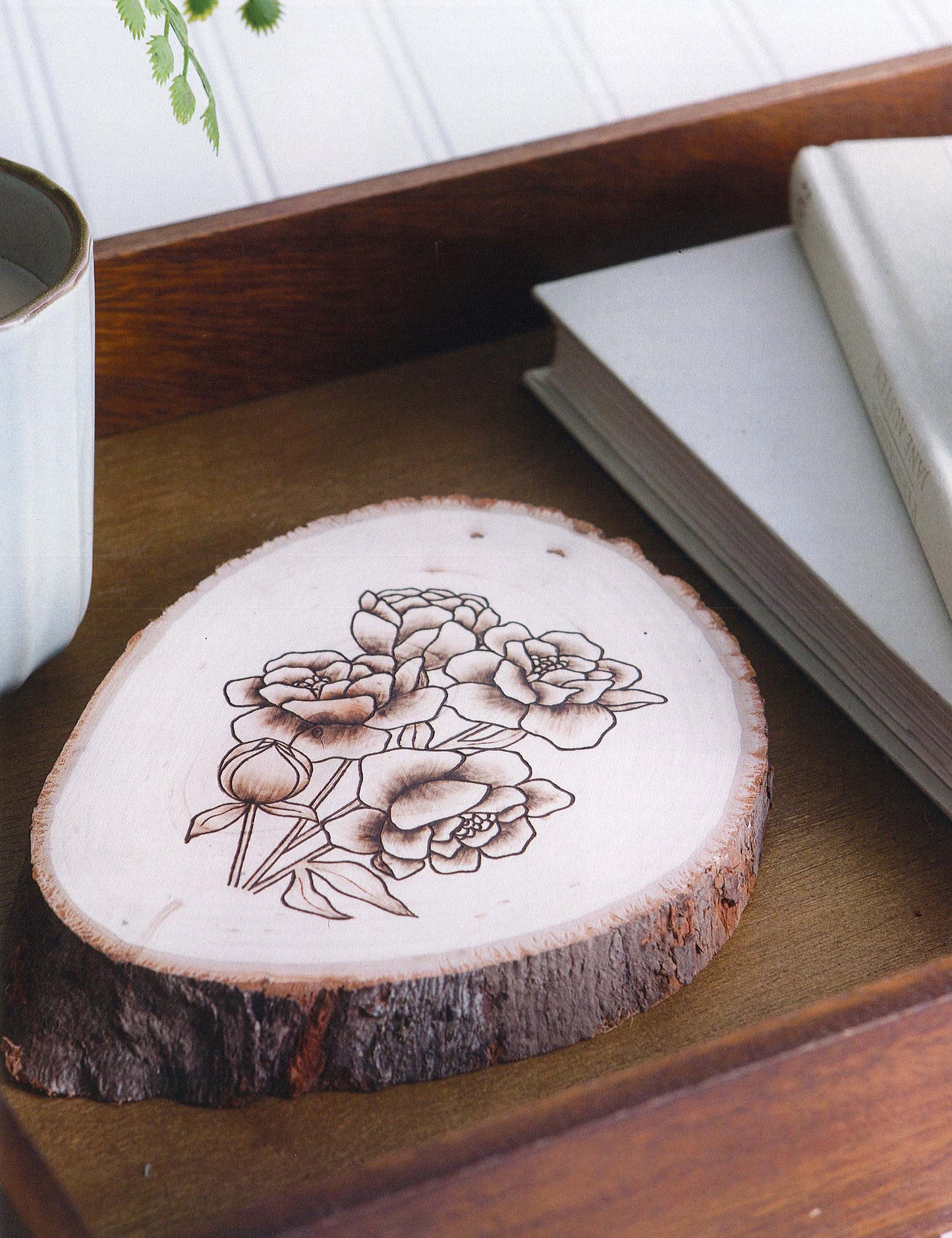 Modern Woodburning 2 by Leisure Arts features a wood slice with an intricate floral design and natural bark edge, ideal for beginner pyrography enthusiasts. Displayed on a tray beside a white cup and two closed books.