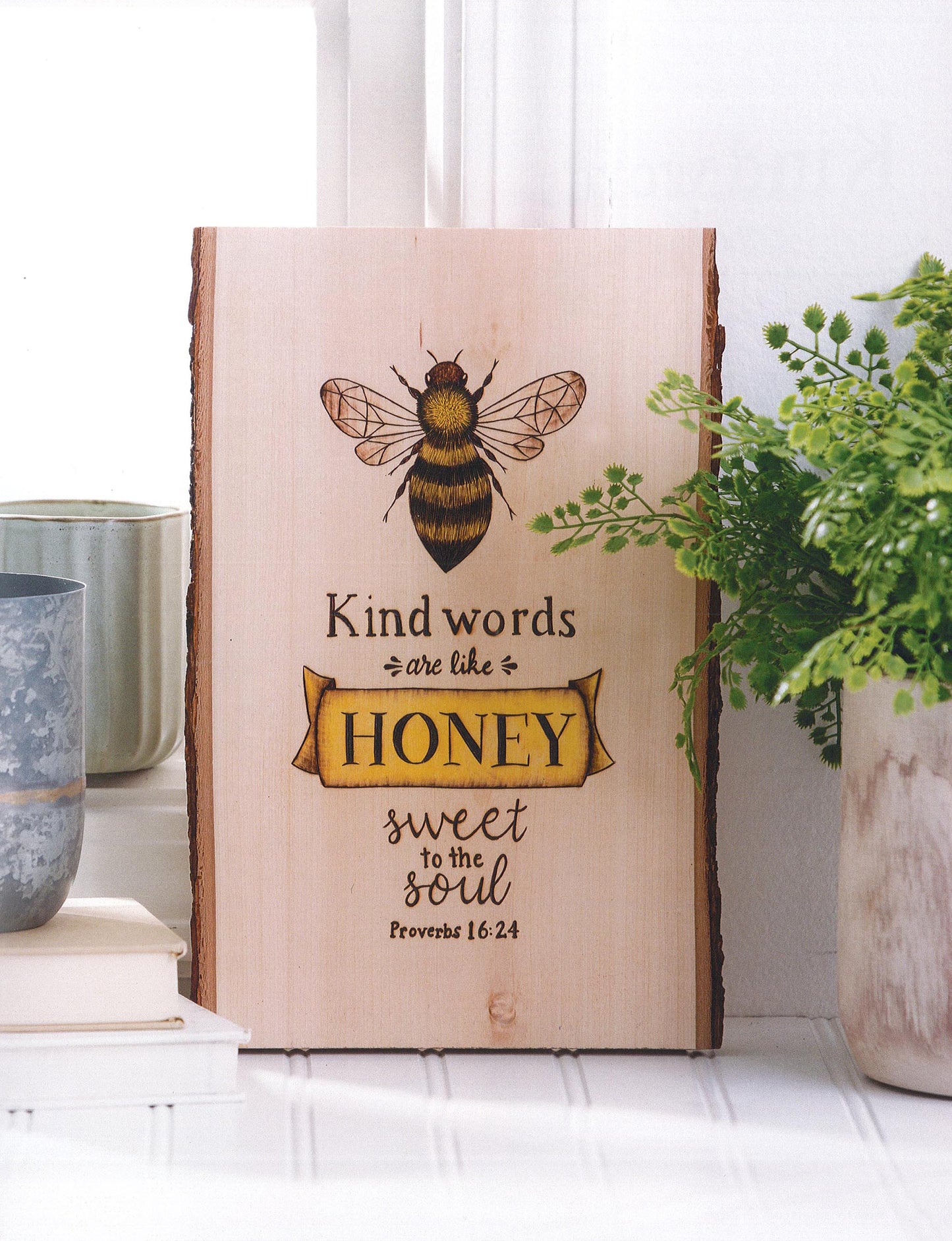 The Modern Woodburning 2 by Leisure Arts is a perfect beginner pyrography project—a wooden sign with a bee, Proverbs 16:24, and the phrase "Kind words are like honey," surrounded by plants and a candle on a windowsill.