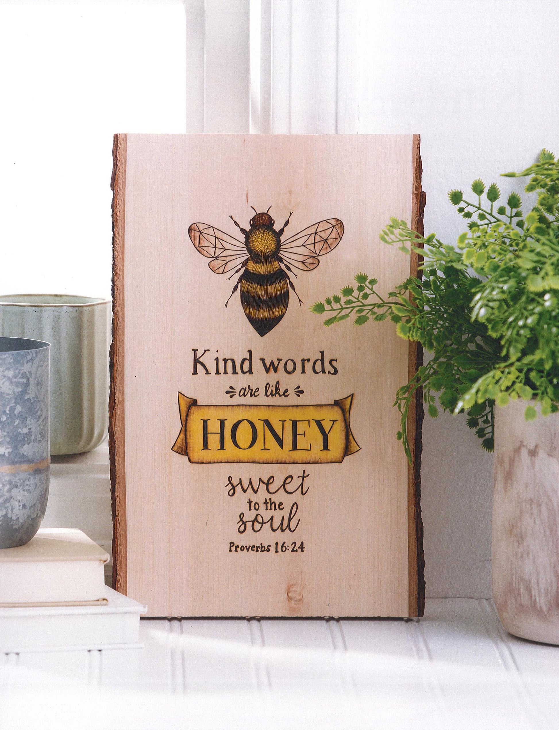 The Modern Woodburning 2 by Leisure Arts is a perfect beginner pyrography project—a wooden sign with a bee, Proverbs 16:24, and the phrase "Kind words are like honey," surrounded by plants and a candle on a windowsill.