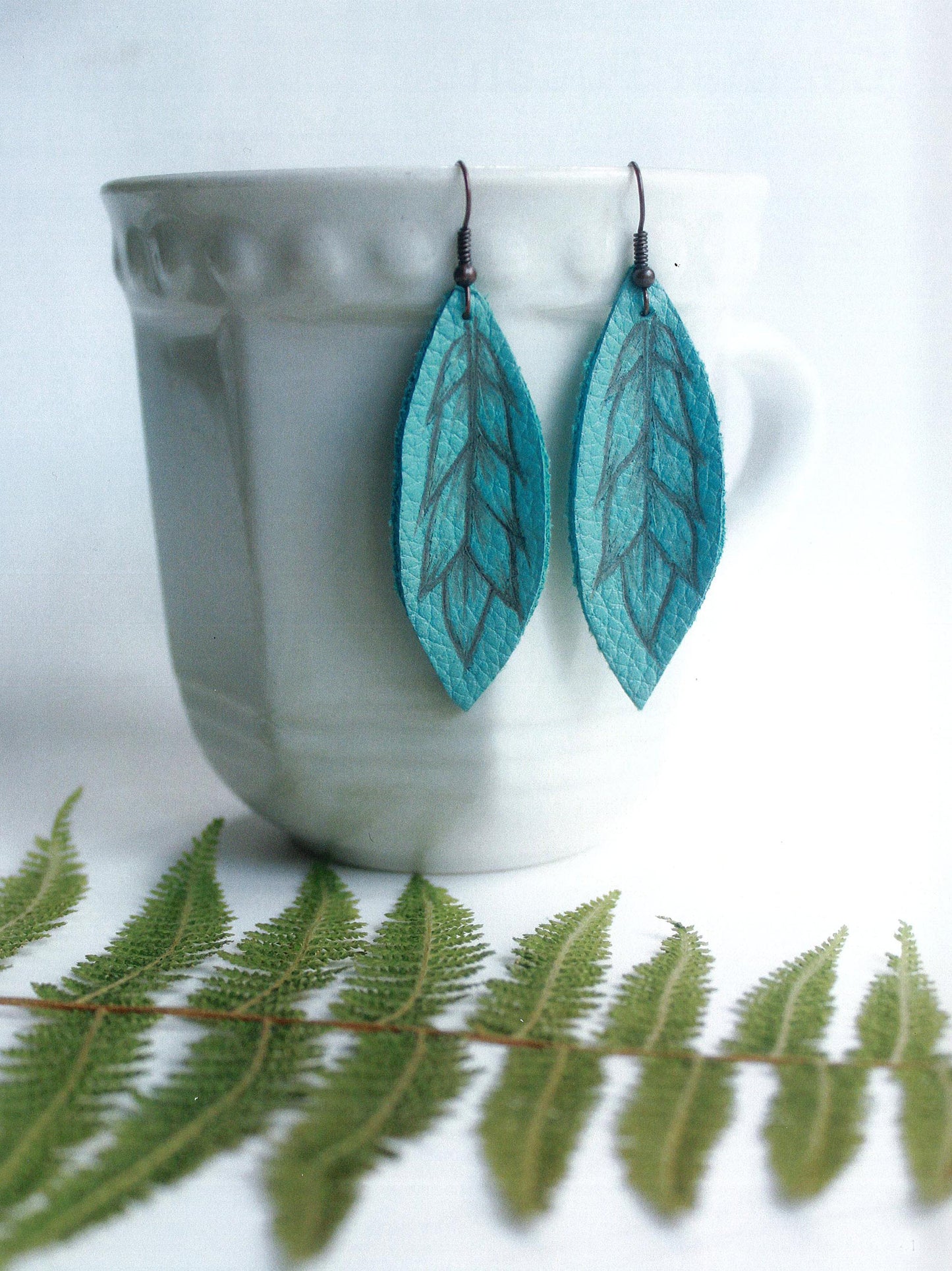 A white ceramic cup displays two teal, leaf-shaped earrings from Leisure Arts’ Modern Woodburning 2—ideal for beginner pyrography. In front, a green fern rests on a white surface.