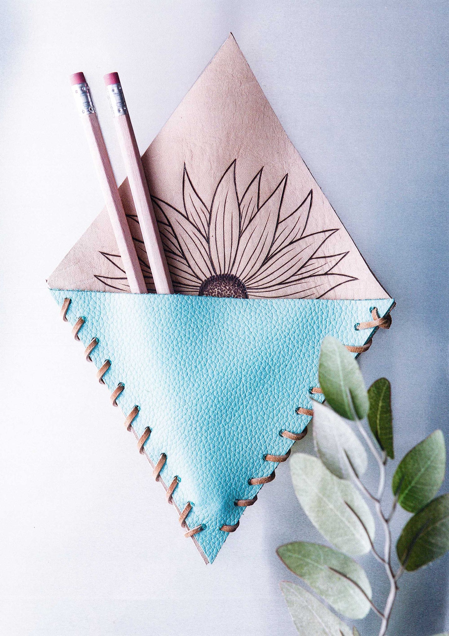 The Modern Woodburning 2 pouch by Leisure Arts is handmade with blue faux leather and tan fabric, showcasing a sunflower inspired by beginner pyrography projects. It holds two pink-eraser pencils and features a leafy branch in the lower right corner.