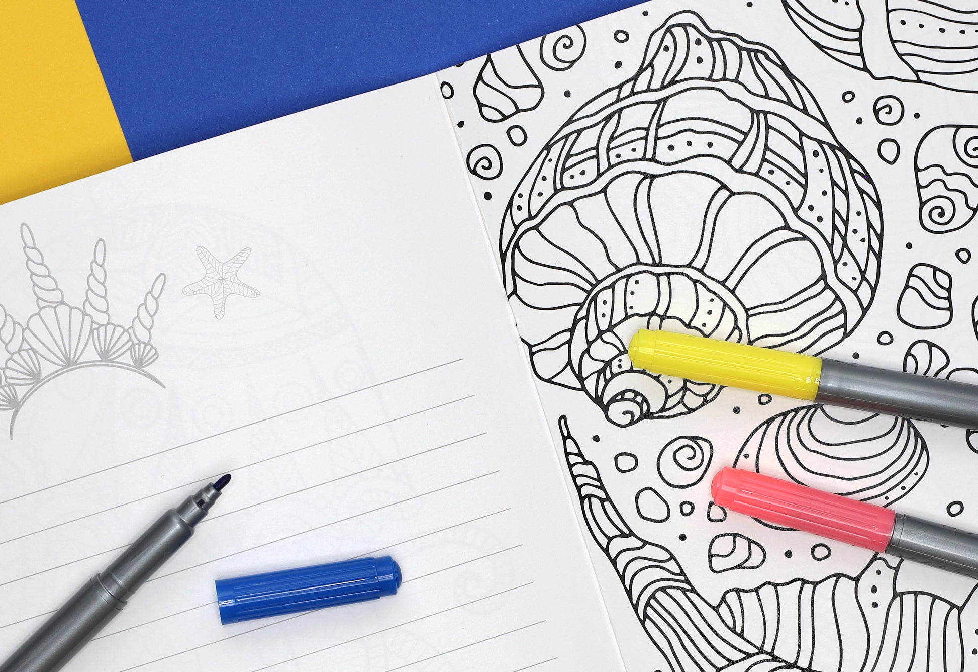 An open Mermaid at Heart Coloring Book by Leisure Arts displays one of 32 seashell designs being colored with yellow, pink, and blue markers. Nearby, a blank lined page and an uncapped black pen sit on a blue and yellow background.