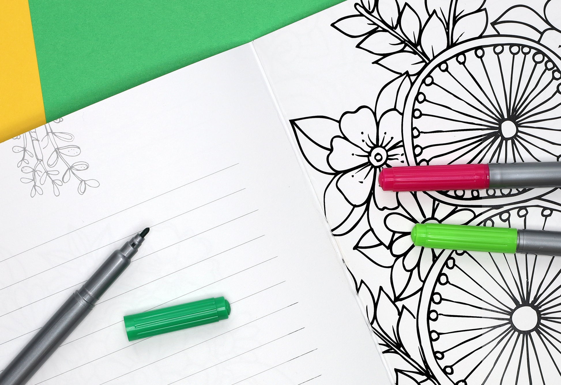 An open lined notebook with a pen and green cap sits beside the Leisure Arts Love Grows Coloring Book, featuring floral designs and three uncapped markers—pink, green, and gray—ready to color all 32 pages. Green and yellow paper brighten the background.