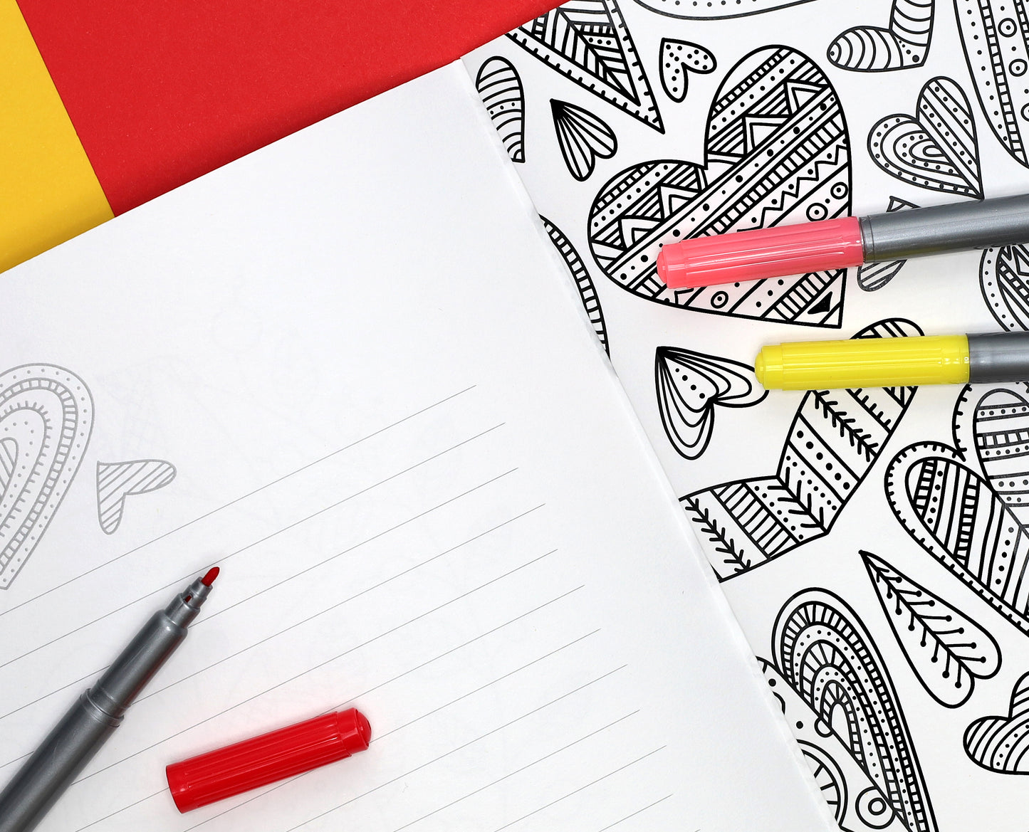 An open notebook with lined pages sits next to a Leisure Arts Grateful Hearts Coloring Book page filled with black-and-white heart designs. Two markers, one red and one yellow, rest on the coloring page; a red marker cap is nearby.