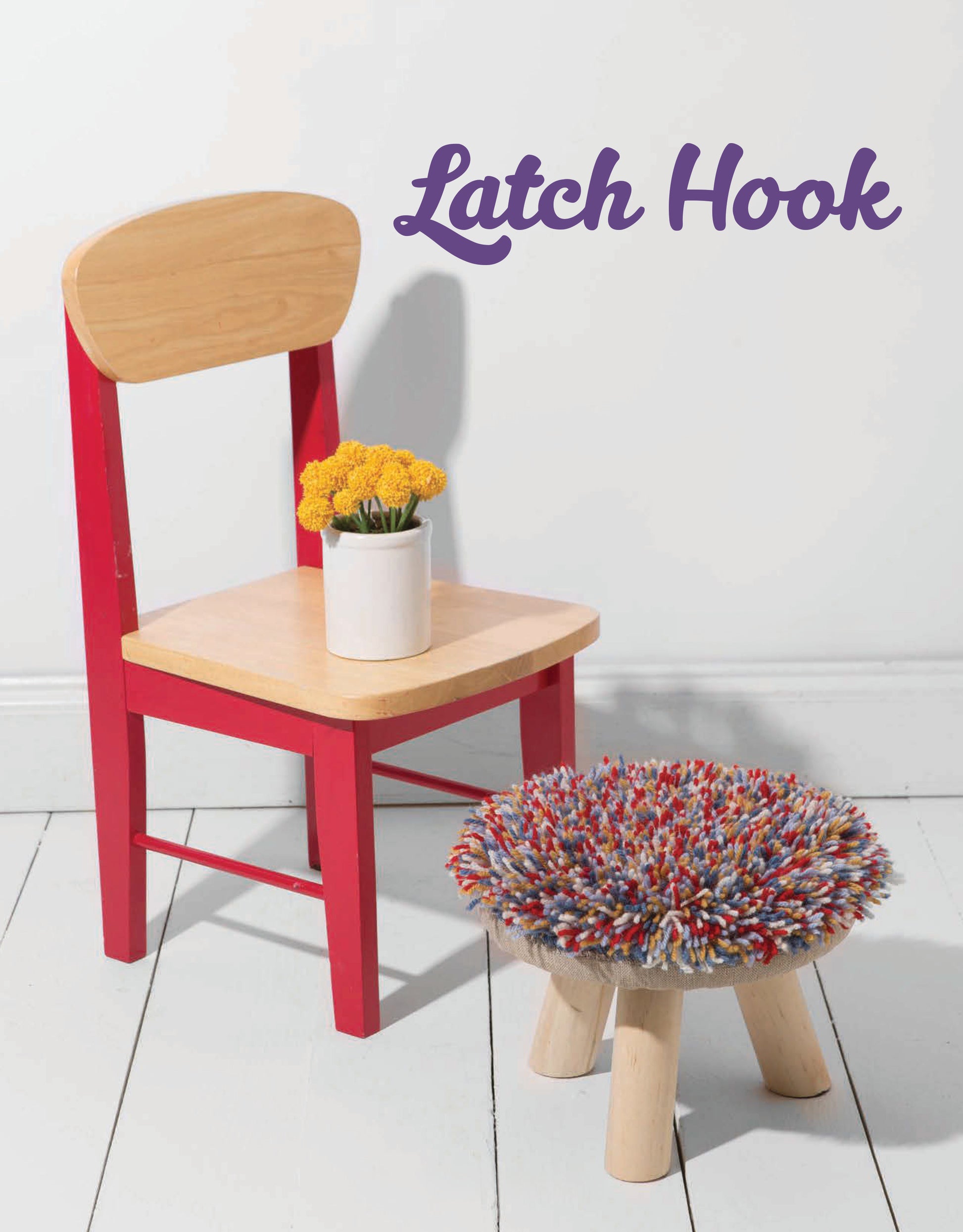A small wooden chair with red legs holds a white vase of yellow flowers. Next to it is a colorful latch hook stool. On the wall behind them, “Latch Hook—12 Projects for the Modern Maker” by Leisure Arts is written.