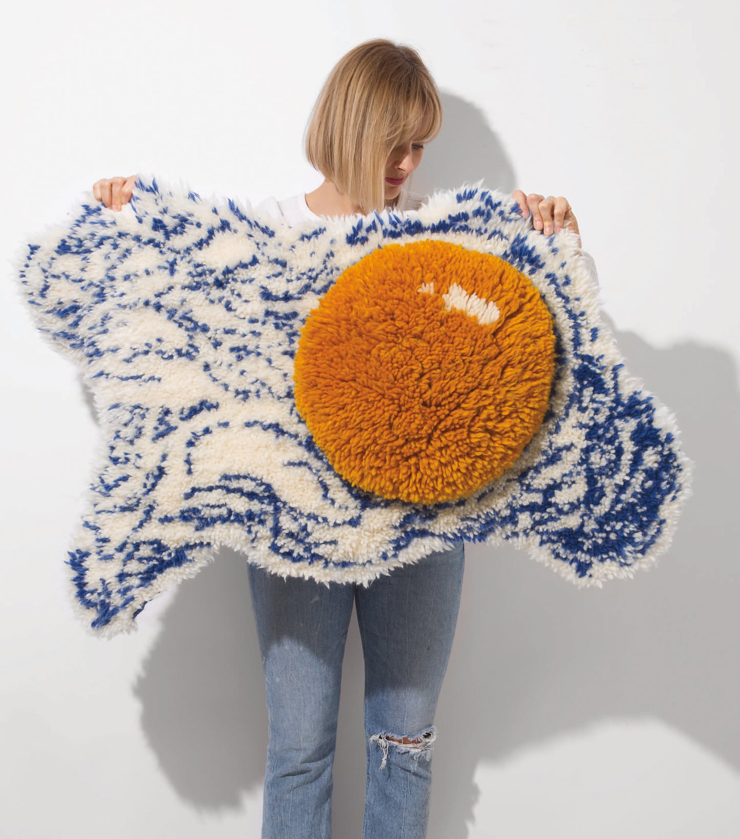 A person with blonde hair holds Leisure Arts' Latch Hook—12 Projects for the Modern Maker Digital Download, featuring a playful fried egg rug design with a bold orange yolk and blue-and-white patterns—an eye-catching home accent.