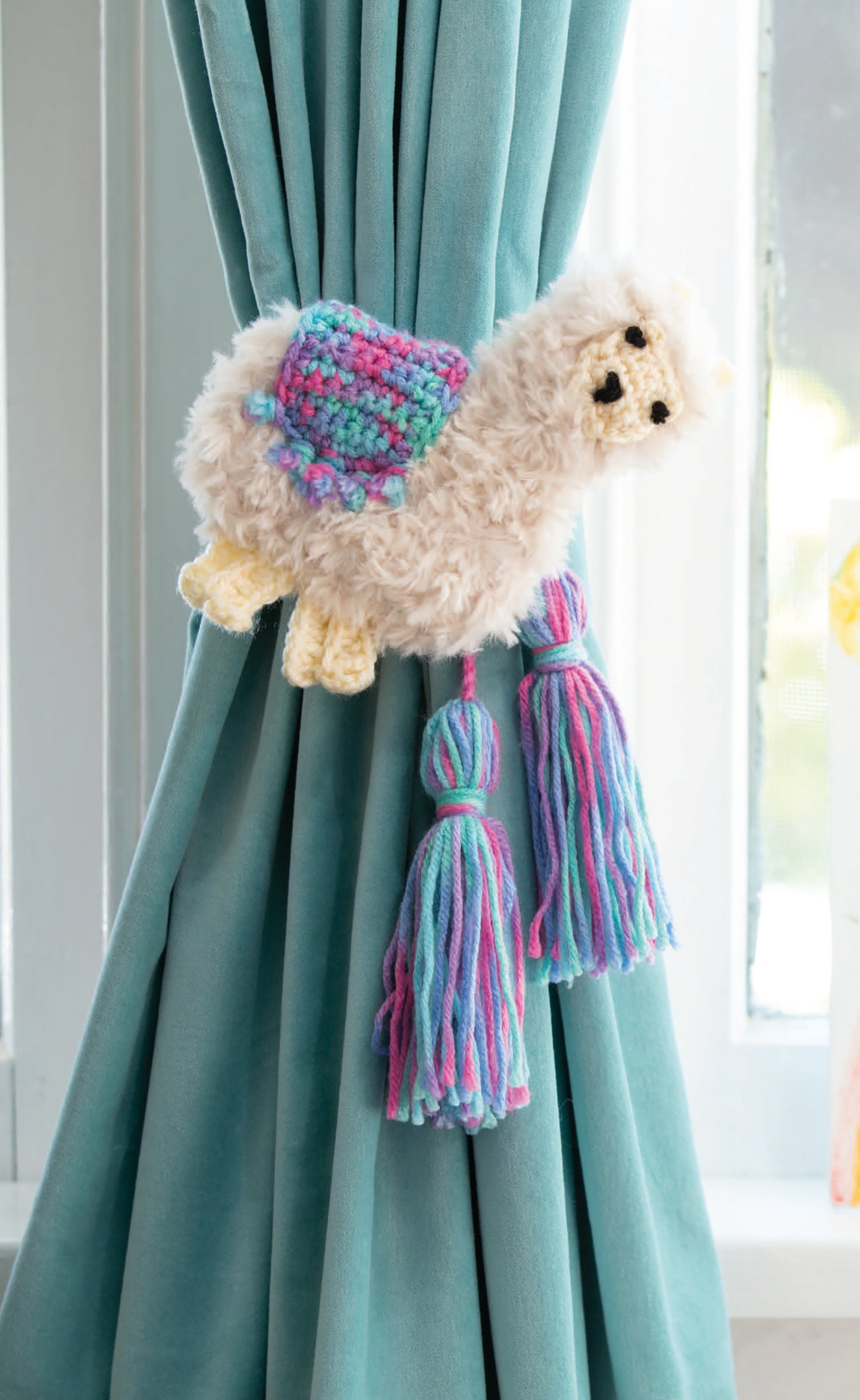 The Leisure Arts Huggable Tie Backs feature a fluffy llama shape with a multicolored crochet saddle and two pastel yarn tassels, adding whimsical charm while stylishly holding back your teal curtain near the window.
