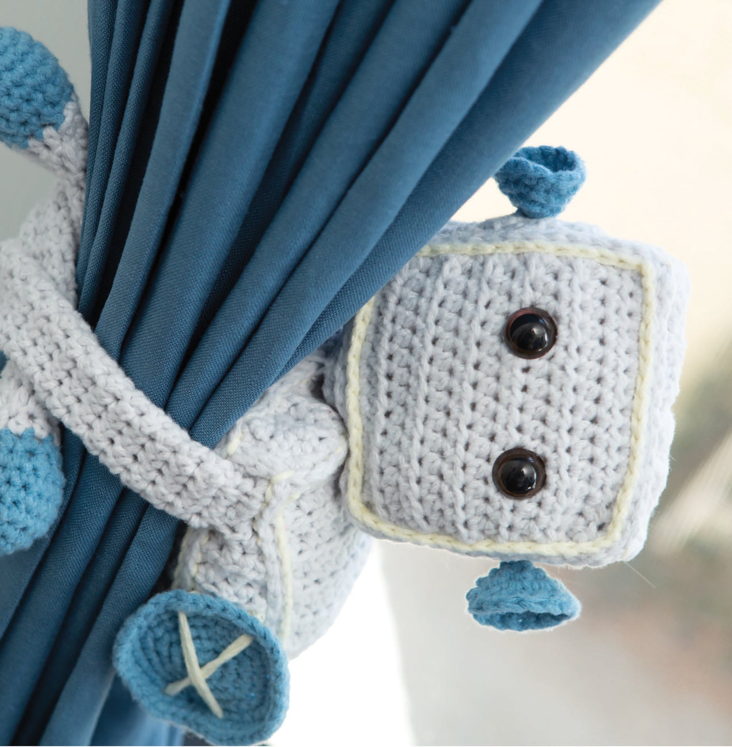 The Huggable Tie Backs by Leisure Arts are crocheted robot curtain holders with button eyes and blue accents, wrapping neatly around your curtain—a cute addition to your home decor and a great beginner project for tie backs.