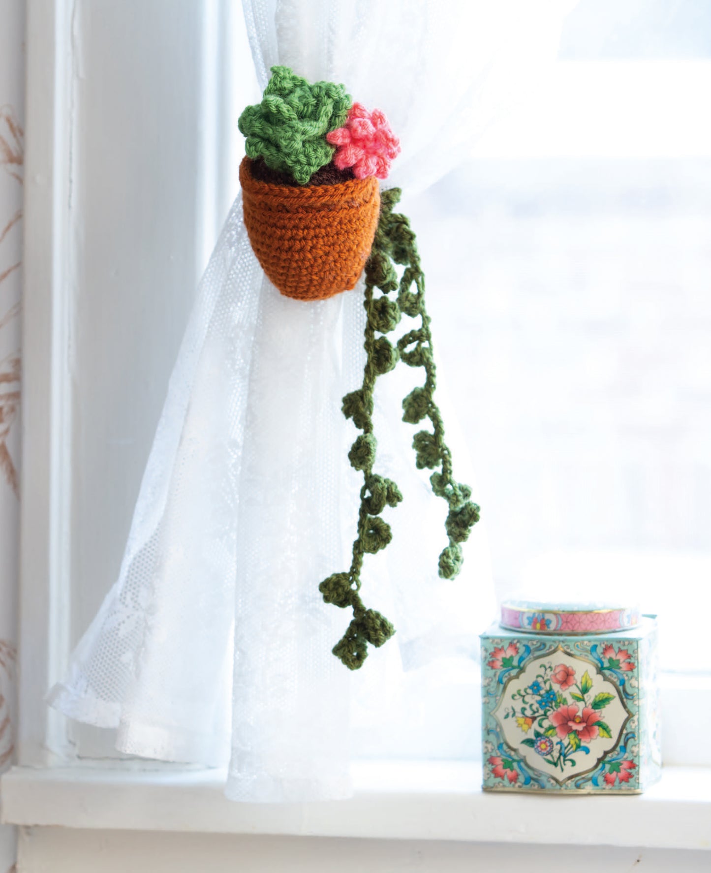 The Huggable Tie Backs by Leisure Arts, featuring a crocheted green-leaf and pink-flower design, make charming curtain tie backs for sheer white curtains—perfect for home decor or beginner projects. A floral tin accents the windowsill.