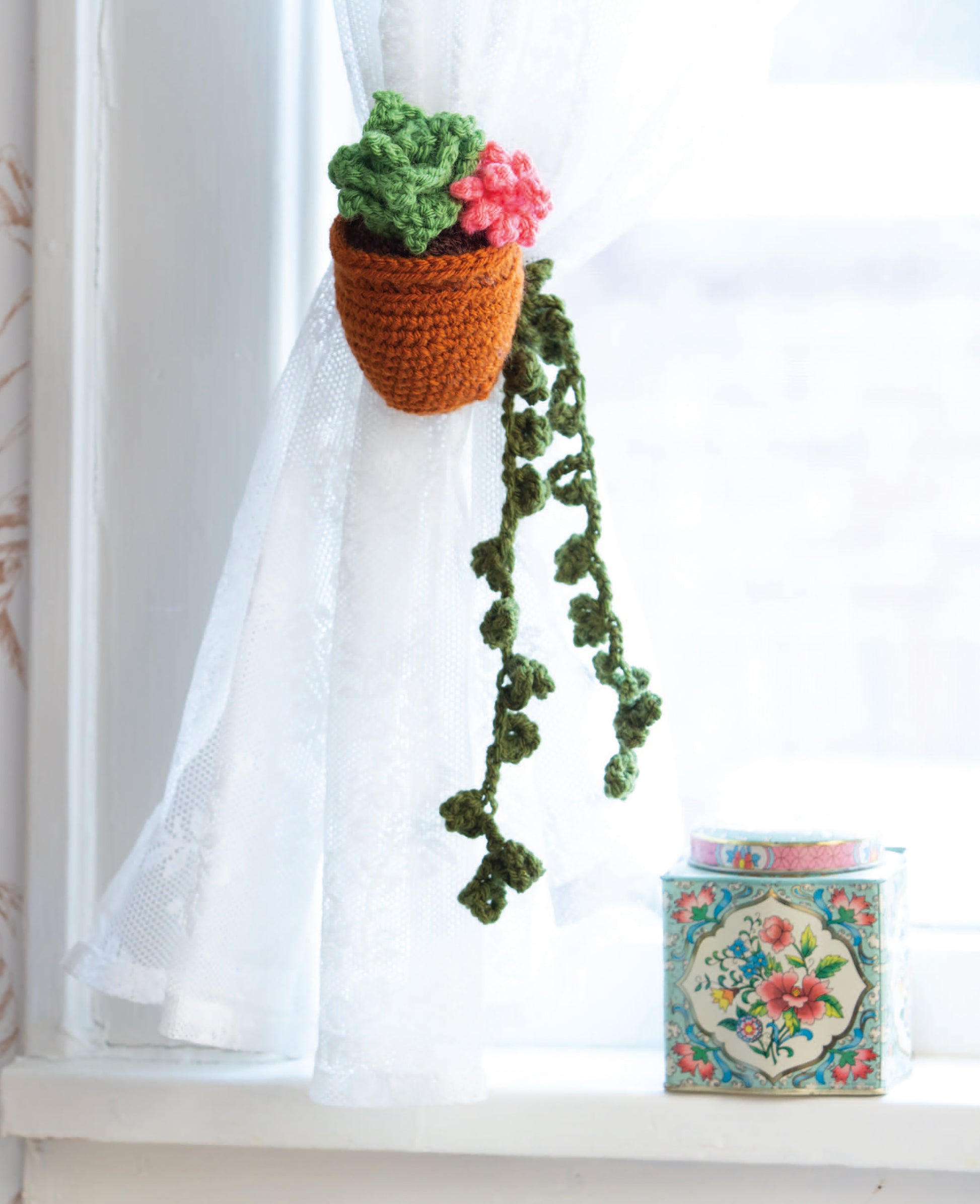 The Huggable Tie Backs by Leisure Arts, featuring a crocheted green-leaf and pink-flower design, make charming curtain tie backs for sheer white curtains—perfect for home decor or beginner projects. A floral tin accents the windowsill.