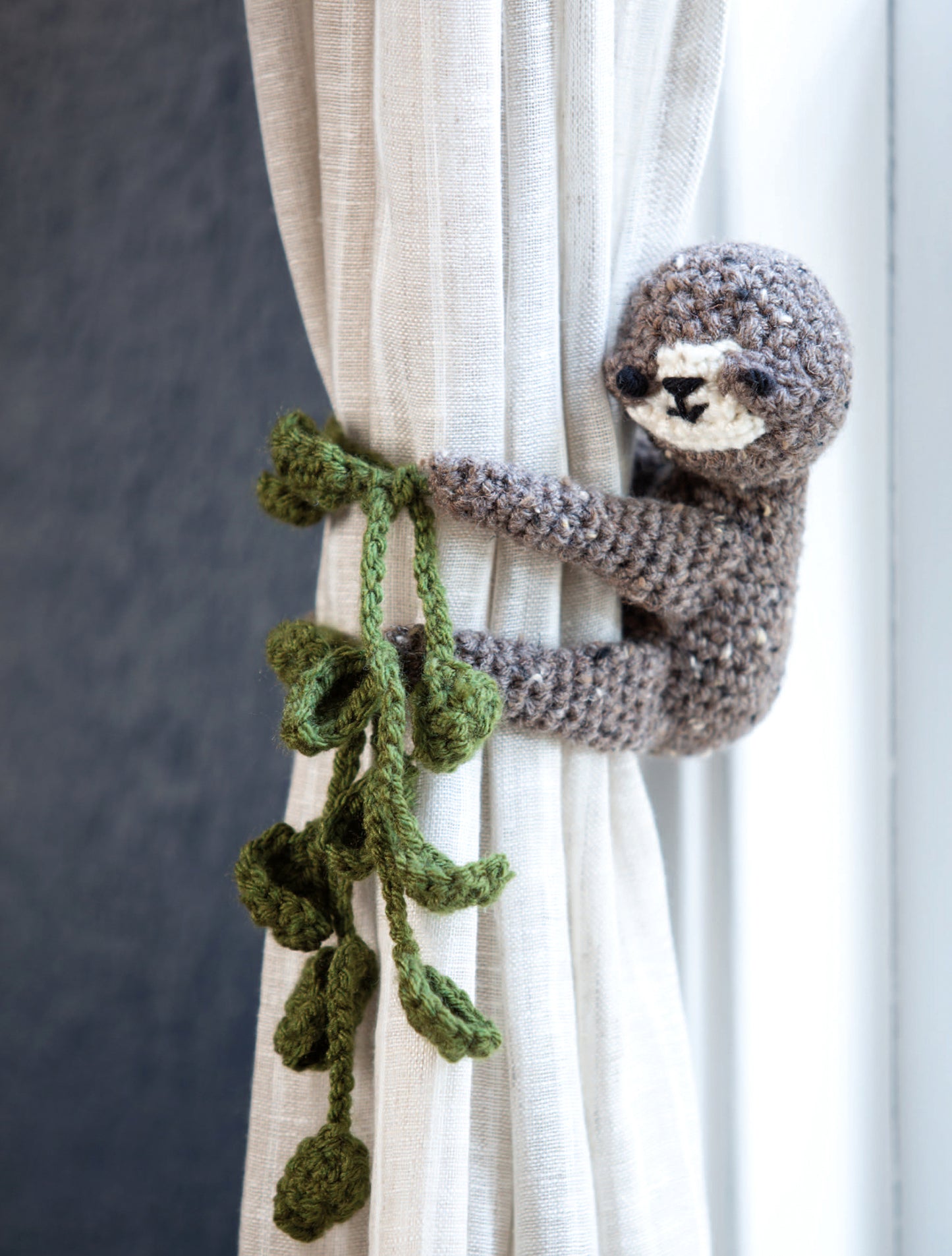 The Leisure Arts Huggable Tie Backs—a crocheted sloth with green yarn leaves—wraps around a white curtain, holding it open. It’s a whimsical home decor accent and an ideal beginner-friendly project for charming curtain tiebacks.