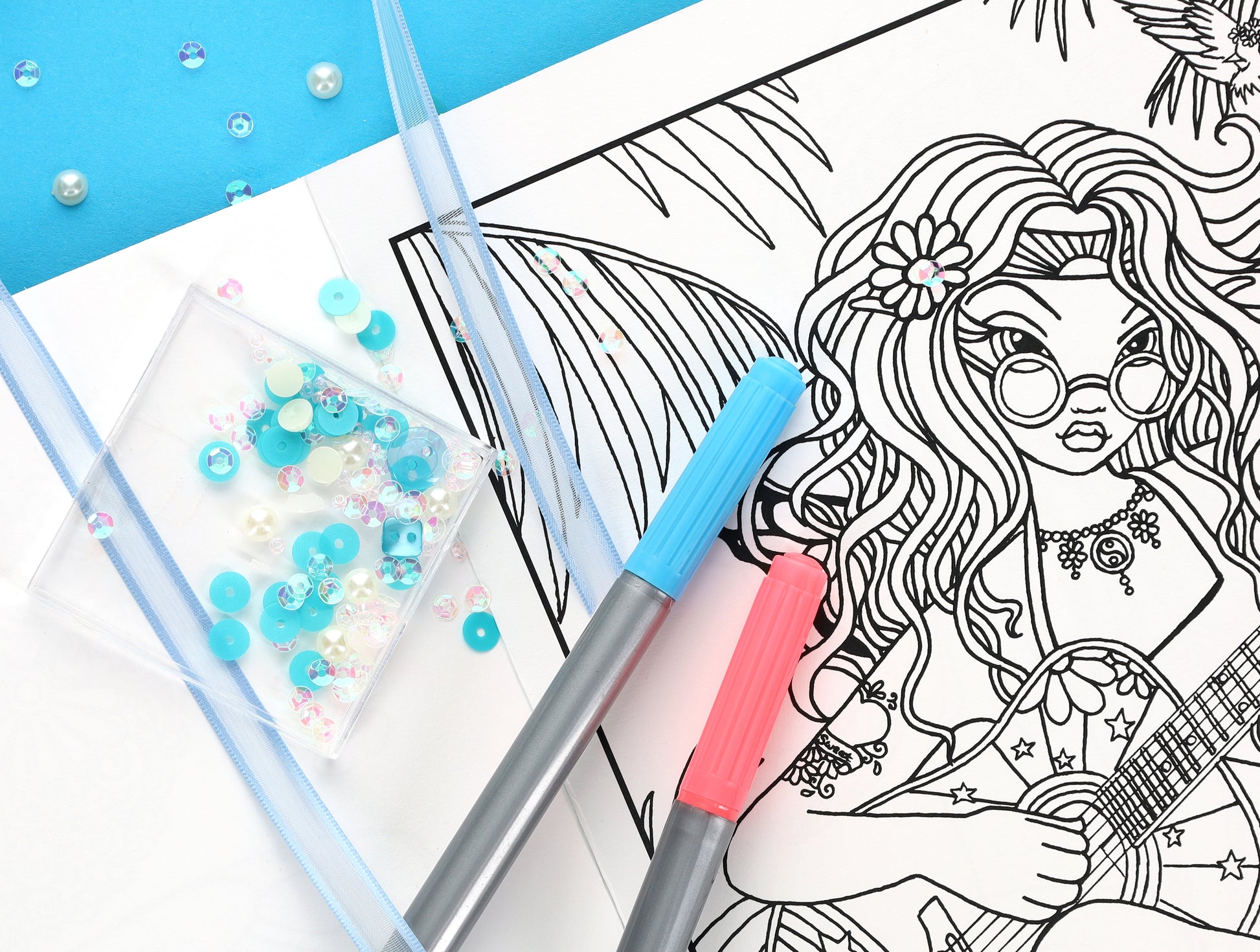 A coloring page from the Leisure Arts Good Vibes—24 Designs; Myka Jelina Coloring Collection shows a girl with flowers in her hair playing guitar, with blue and pink markers, blue ribbon, and sequins on a bright blue background.