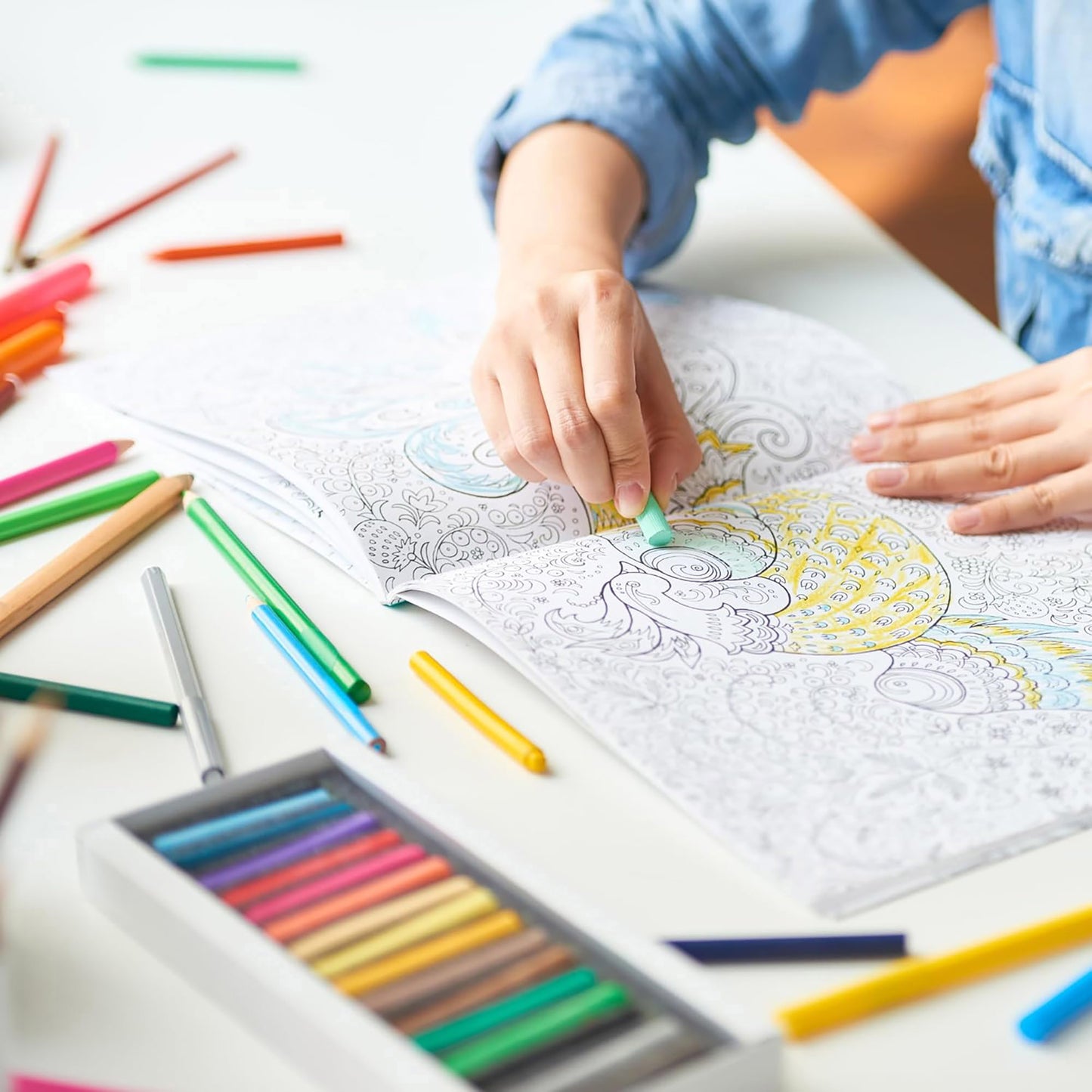 A person colors the Be Yourself Coloring Book—24 Designs from the Myka Jelina Coloring Collection by Leisure Arts with colored pencils and pastels on a white table, surrounded by more Leisure Arts supplies; only their hands and torso are visible.