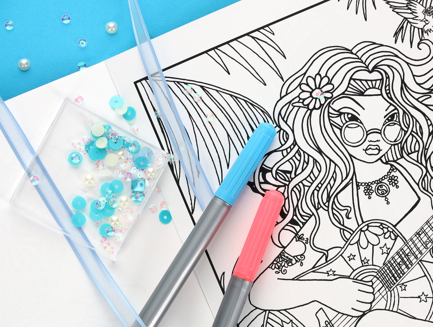 A partially colored-in Good Vibes—32 Designs; Myka Jelina Coloring Collection page by Leisure Arts shows a girl with glasses playing guitar. Blue and pink markers, blue and white sequins, and a blue ribbon sit on the page.