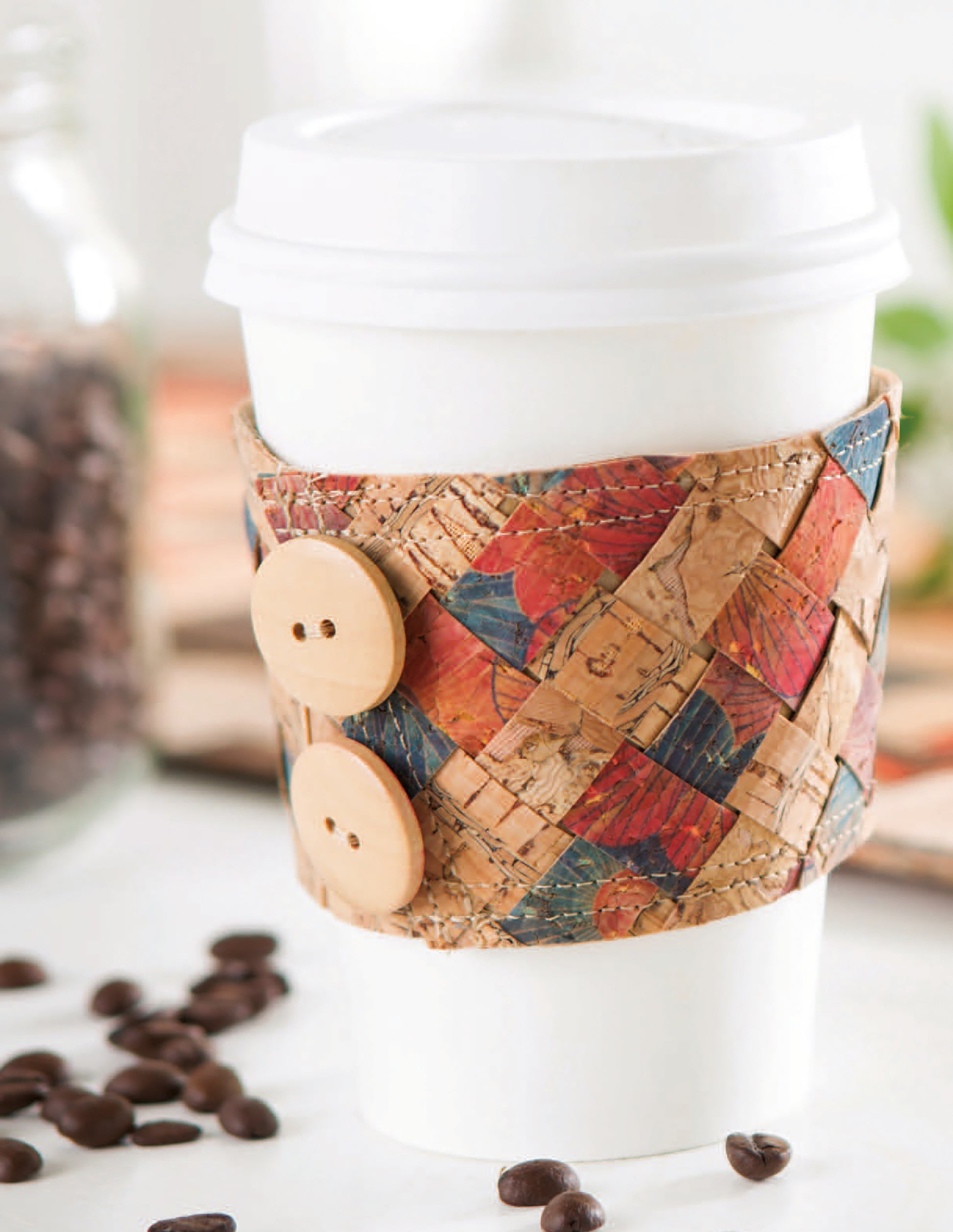 The image shows a white disposable coffee cup with a colorful, woven cork sleeve accented by two large wooden buttons—a project idea from Cork Creations—20+ Modern Sew & No-Sew Cork Creations Digital Download by Leisure Arts. Coffee beans and a glass jar add flair.