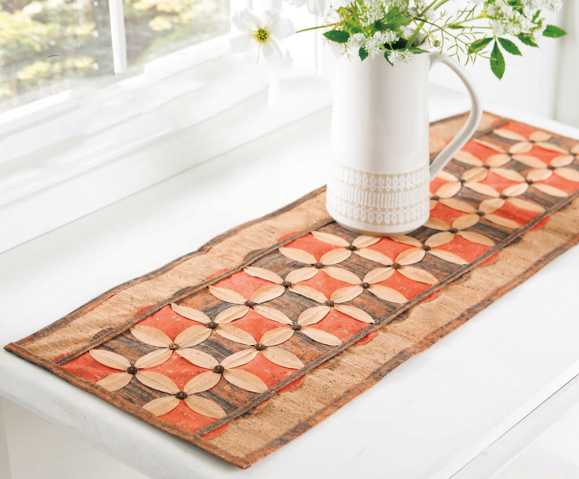 A white ceramic pitcher with flowers rests on a white table, adorned with a quilted runner. Add charm to your space or sewing projects with Cork Creations—20+ Modern Sew & No-Sew Patterns, a digital download from Leisure Arts. Sunlight pours in nearby.