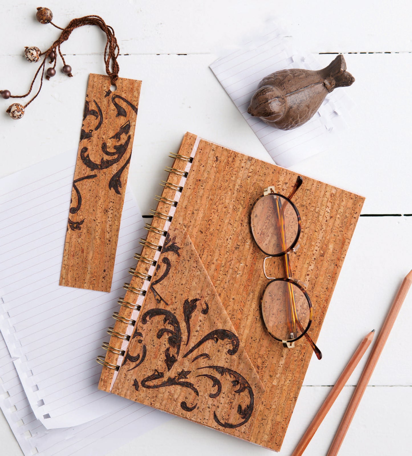 On a white wooden surface: Leisure Arts' Cork Creations digital download, matching bookmark, reading glasses, 2 pencils, a wooden bird figurine, and lined paper—ideal for beginner crafts with 20+ modern cork projects.