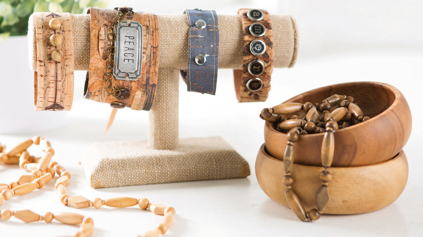 A display featuring beaded bracelets, a necklace, and cork wristbands with charms—crafted for cork enthusiasts—highlights Leisure Arts' "Cork Creations—20+ Modern Sew & No-Sew Cork Creations" digital download, arranged on a beige stand and in wooden bowls.