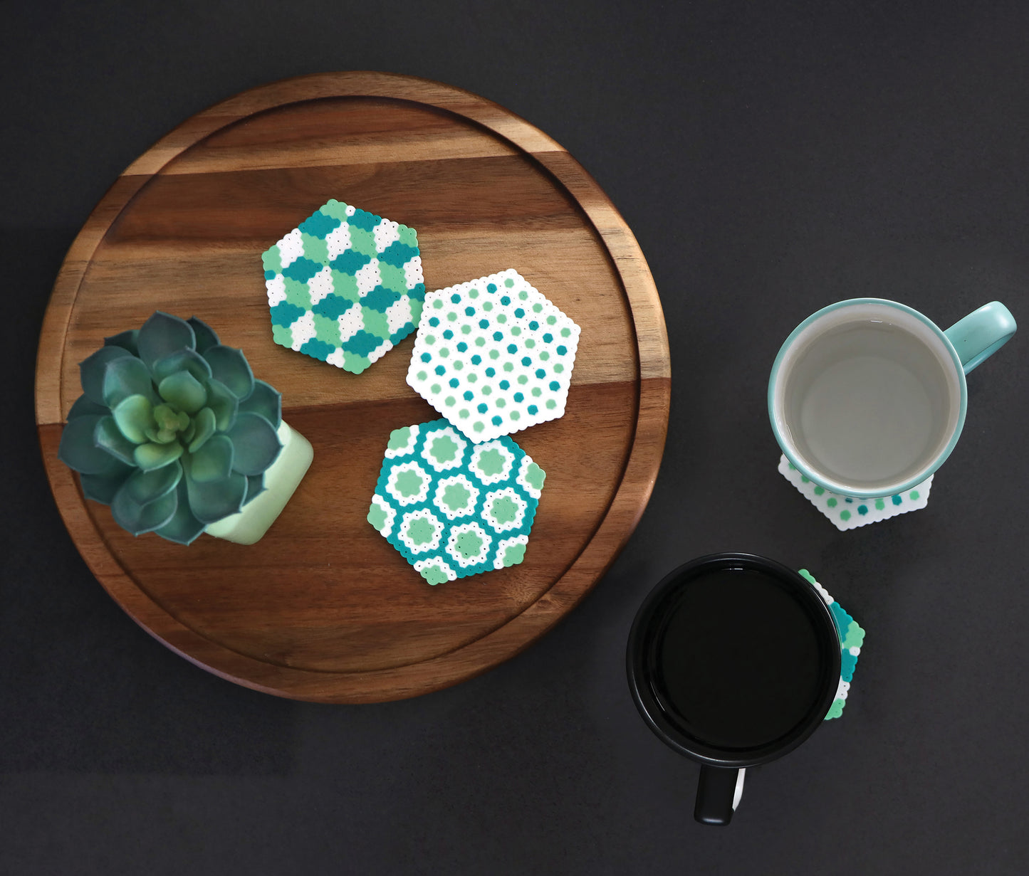On a round wooden tray, a small succulent and three hexagonal coasters—crafted from the "Melt and Make Bead Crafts Digital Download" by Leisure Arts—are arranged. Nearby, two mugs rest on coasters against a dark background.