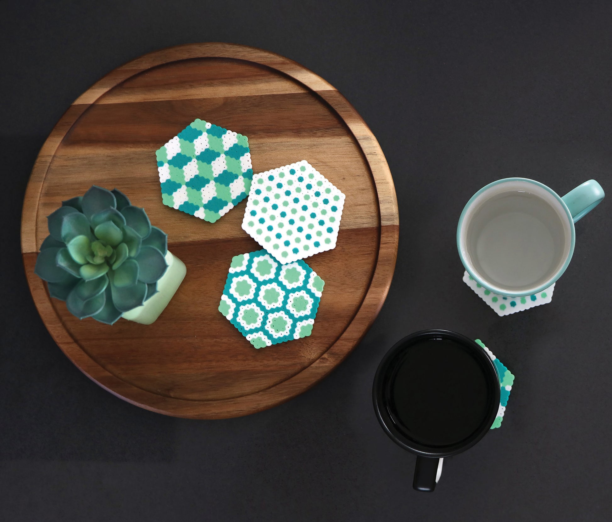 On a round wooden tray, a small succulent and three hexagonal coasters—crafted from the "Melt and Make Bead Crafts Digital Download" by Leisure Arts—are arranged. Nearby, two mugs rest on coasters against a dark background.